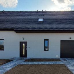 Coloseum Pro sp. z o.o. - Nowoczesny, parterowy dom z garażem, wykończony w jasnych kolorach z ciemną dachówką i stolarką okienną. Uwagę zwraca minimalistyczny styl i kostka brukowa prowadząca do wejścia.