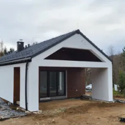 Nowoczesny dom modułowy z białymi ścianami i ciemnym dachem, z otwartą przestrzenią garażową i drewnianymi panelami, w trakcie budowy, widoczne materiały budowlane na ziemi.