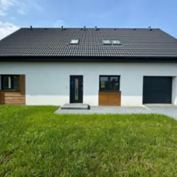 Nowoczesny, parterowy dom z białymi ścianami, ciemną dachówką i dwoma oknami dachowymi, z garażem po prawej stronie i fragmentami elewacji wykończonymi drewnopodobną okładziną. Wejście...