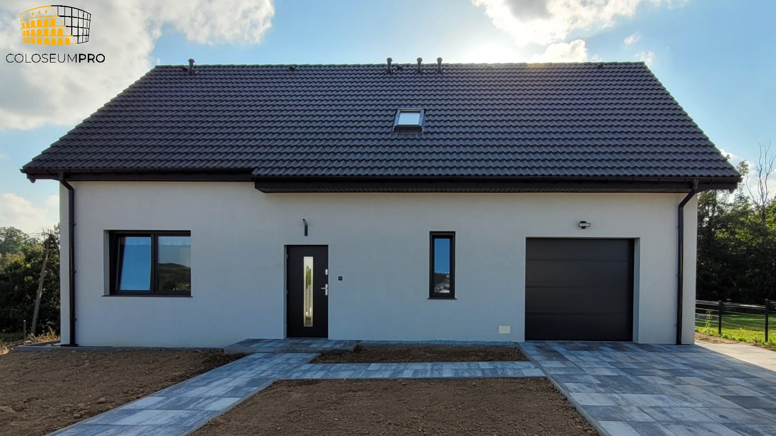 Nowoczesny, parterowy dom z garażem, wykończony w jasnych kolorach z ciemną dachówką i stolarką okienną. Uwagę zwraca minimalistyczny styl i kostka brukowa prowadząca do wejścia.