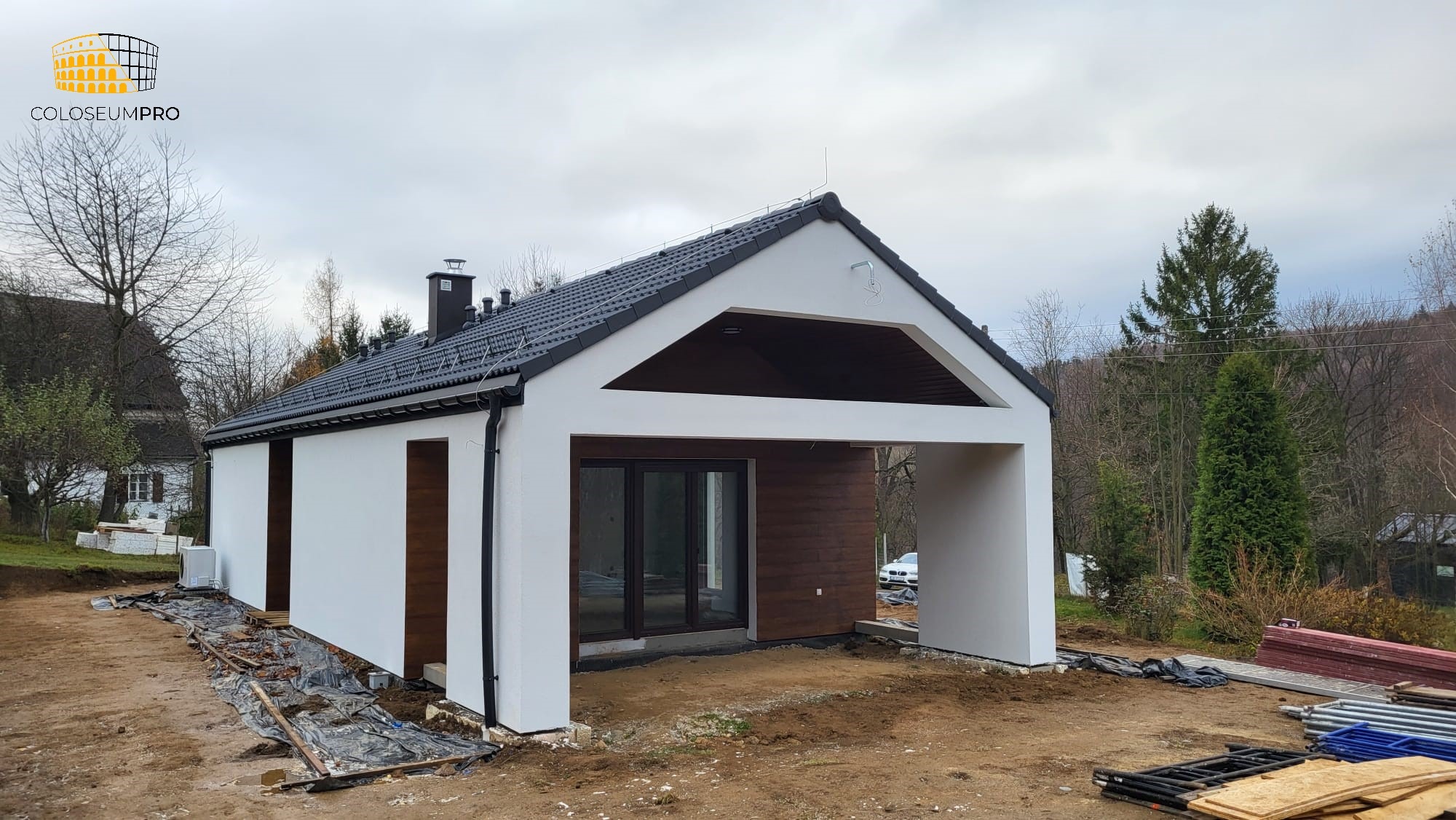 Nowoczesny dom modułowy z białymi ścianami i ciemnym dachem, z otwartą przestrzenią garażową i drewnianymi panelami, w trakcie budowy, widoczne materiały budowlane na ziemi.