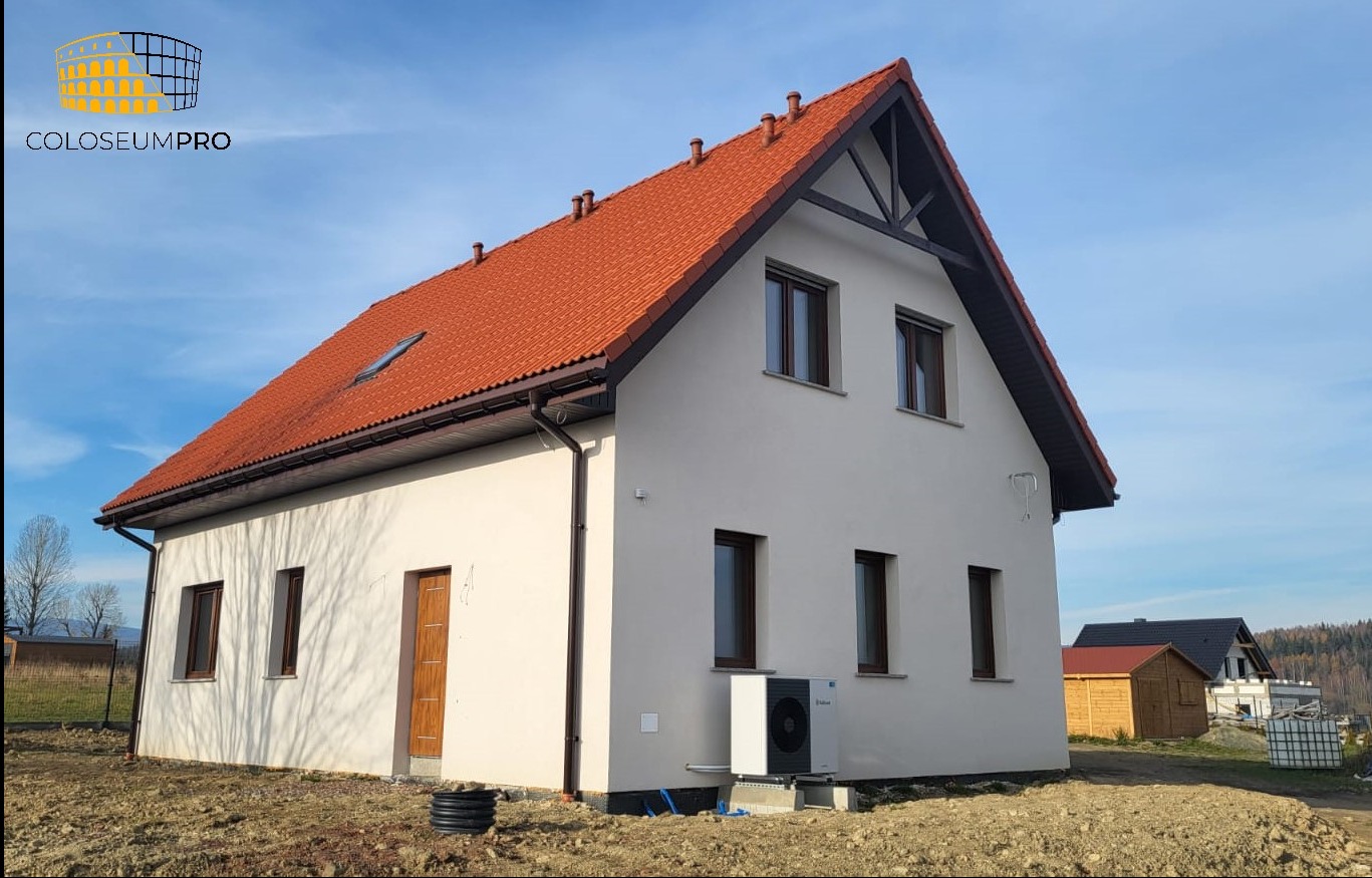 Niedawno wybudowany dom jednorodzinny z jasną elewacją, czerwoną dachówką i zamontowaną pompą ciepła na zewnątrz, widok z boku na tle błękitnego nieba i gór.