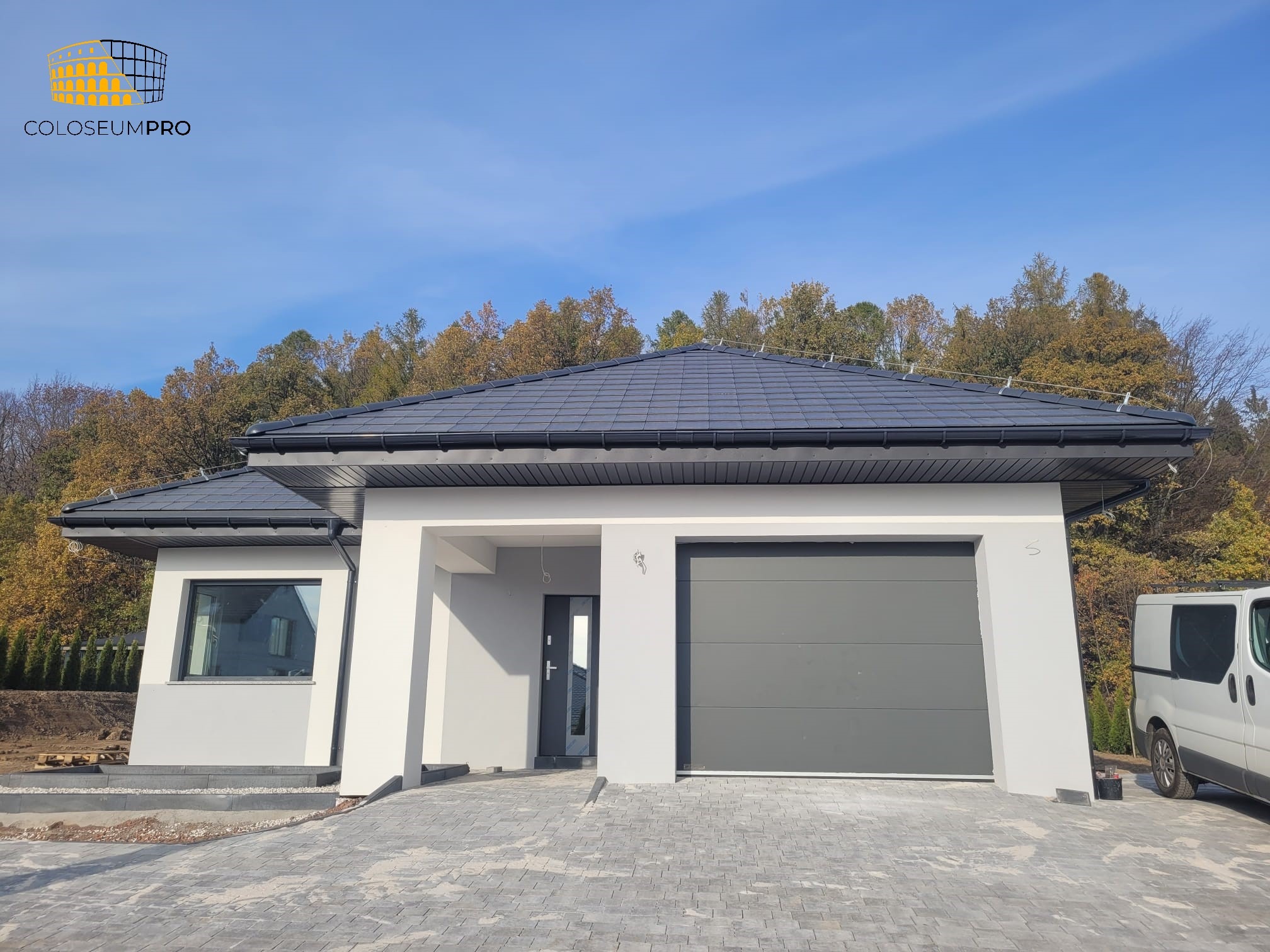 Parterowy budynek z szarą dachówką, grafitową bramą garażową i antracytowymi oknami, wykończony na biało, z kostką brukową na podjeździe i jesiennym lasem w tle.