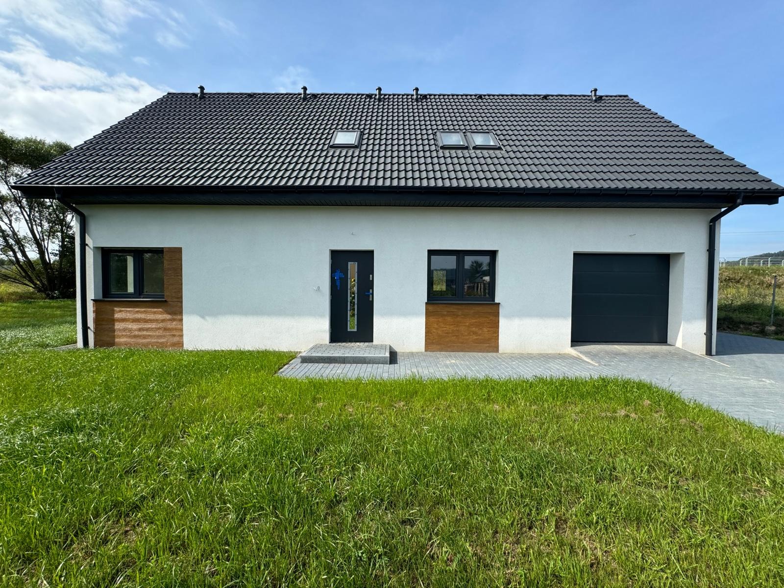 Nowoczesny, parterowy dom z białymi ścianami, ciemną dachówką i dwoma oknami dachowymi, z garażem po prawej stronie i fragmentami elewacji wykończonymi drewnopodobną okładziną. Wejście...