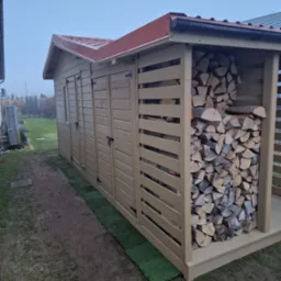 Drewniana konstrukcja z zadaszeniem i miejscem na składowanie drewna, widoczna na tle ogrodu w pochmurny dzień. Konstrukcja pomalowana na jasny kolor.