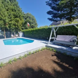 Jasnoszary taras z desek kompozytowych okala basen; obok huśtawka ogrodowa i rabata z posadzonymi roślinami. W tle zieleń drzew i krzewów.