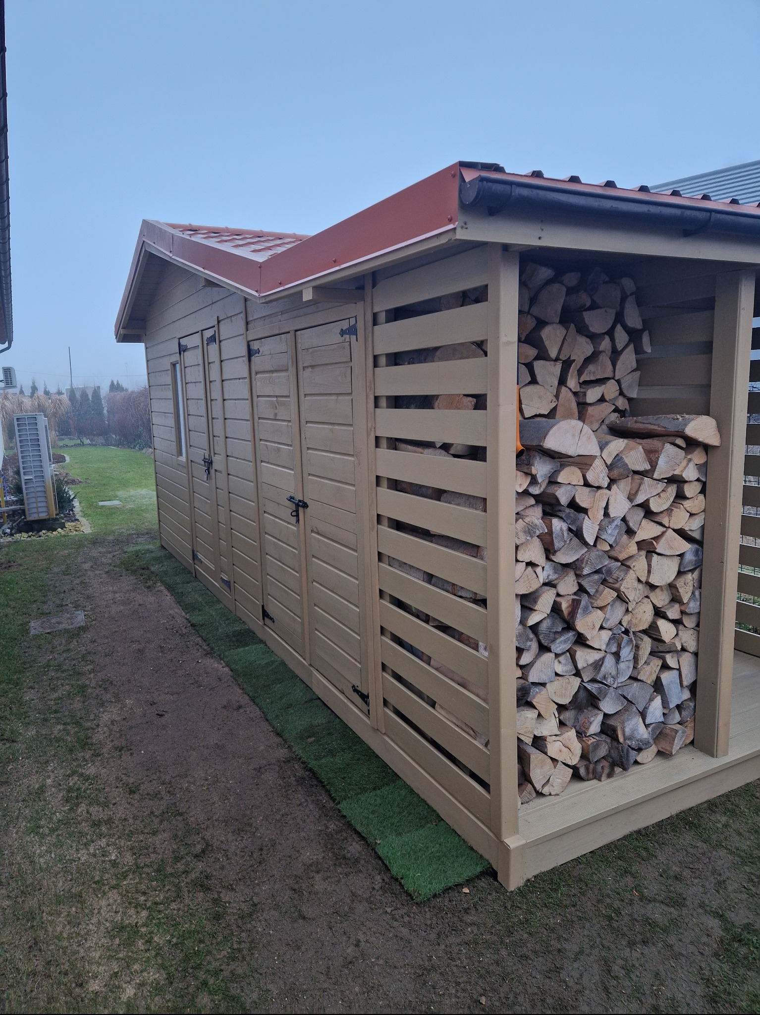 Drewniana konstrukcja z zadaszeniem i miejscem na składowanie drewna, widoczna na tle ogrodu w pochmurny dzień. Konstrukcja pomalowana na jasny kolor.