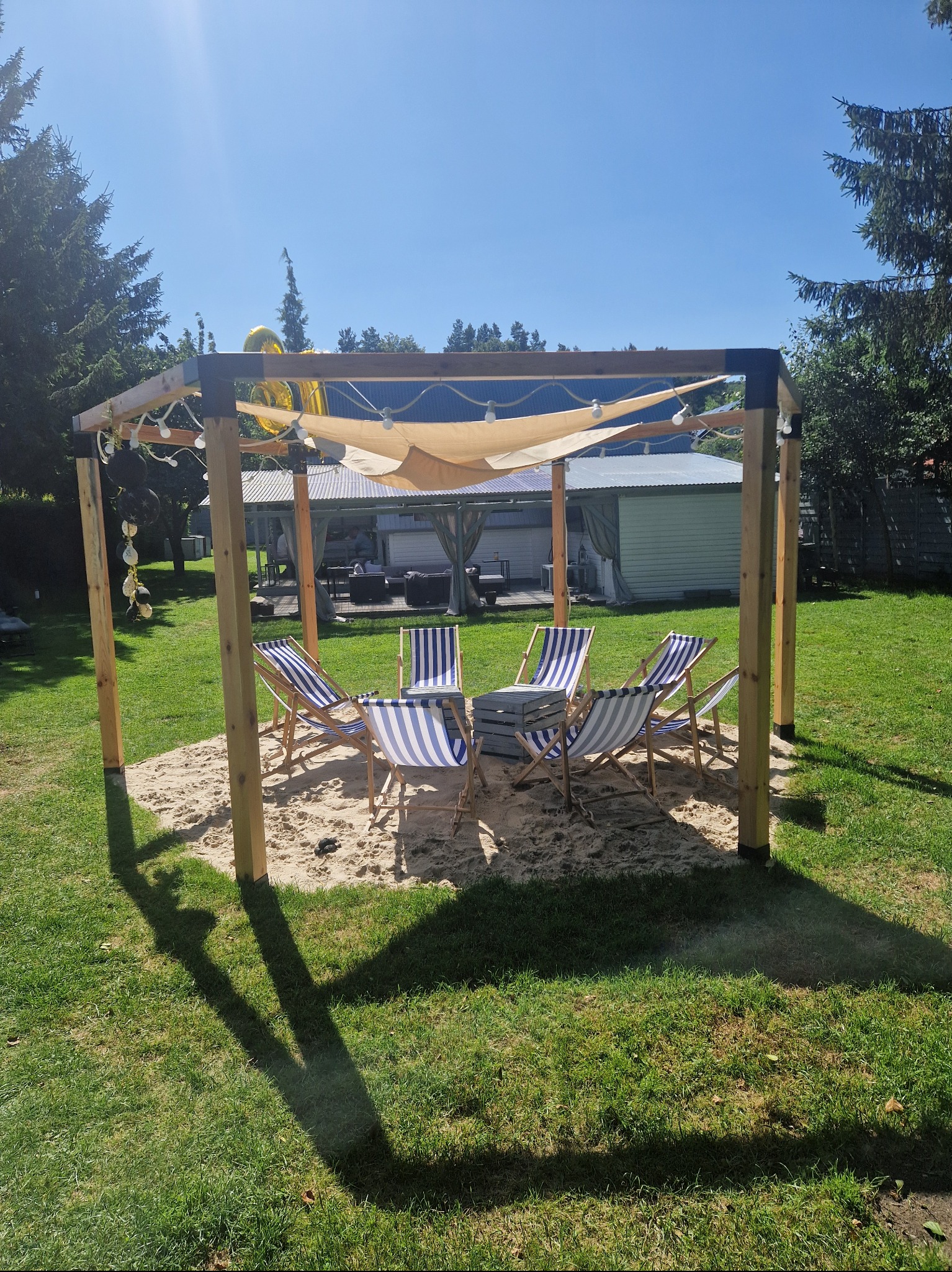 Pergola z jasną płachtą i dekoracjami, otoczona piaskiem i leżakami w paski, tworząca strefę relaksu w ogrodzie. Widoczne drzewa w tle.
