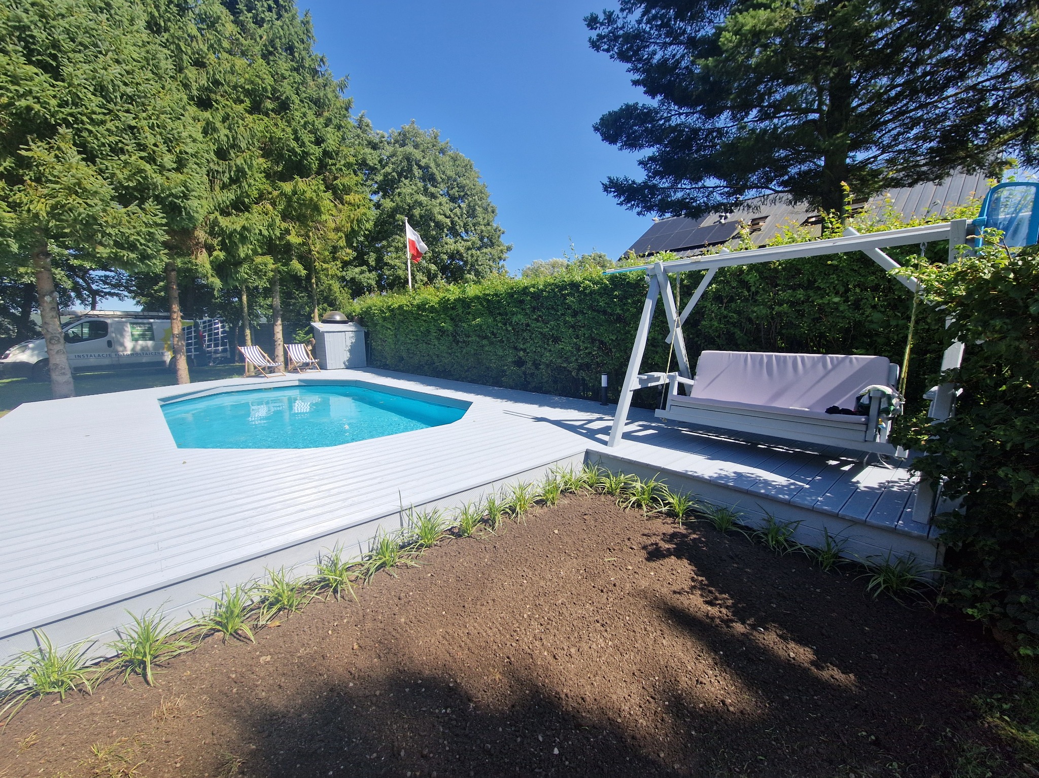 Jasnoszary taras z desek kompozytowych okala basen; obok huśtawka ogrodowa i rabata z posadzonymi roślinami. W tle zieleń drzew i krzewów.