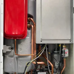 Wnętrze pieca gazowego z czerwonym zbiornikiem wyrównawczym, miedzianymi rurami i szarym panelem. Widoczne liczne przewody i elementy instalacji.