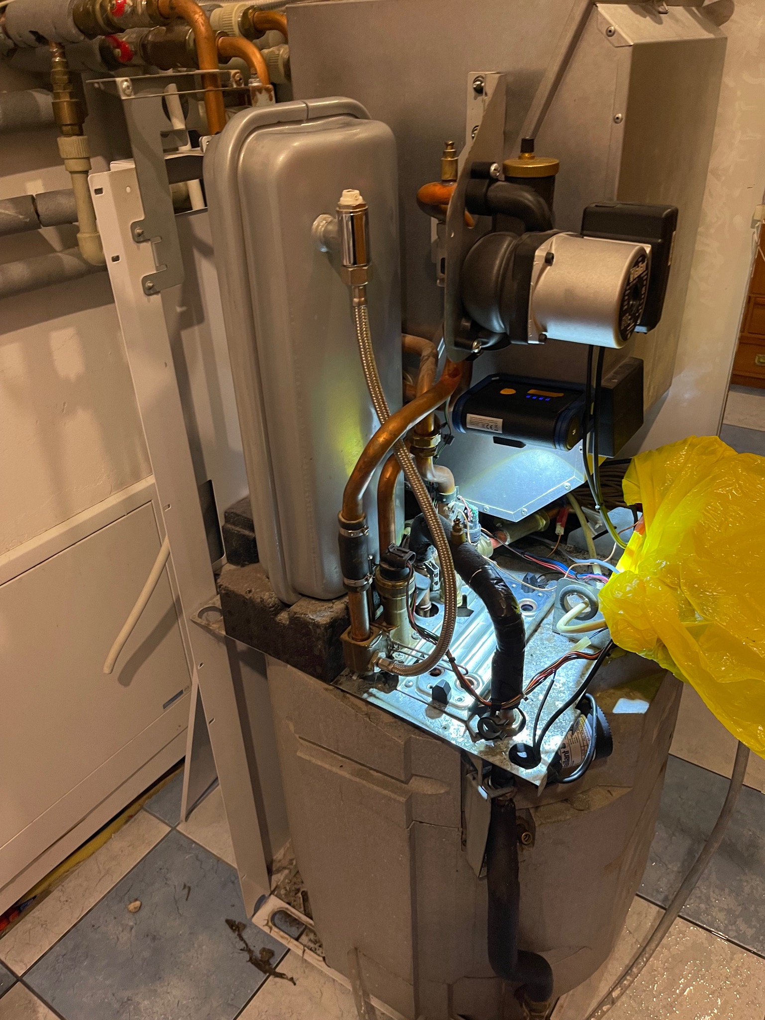 Otwarty piec gazowy z widocznymi elementami instalacji i lampą roboczą, sugerujący serwis lub naprawę. Żółta torba obok.