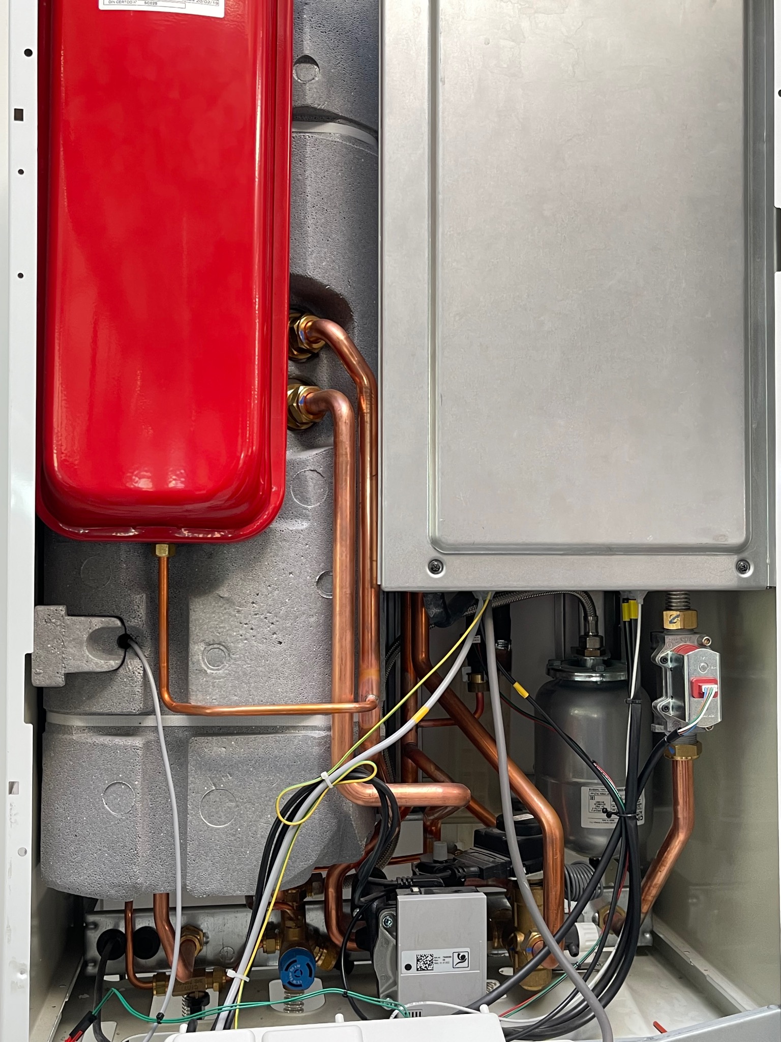 Wnętrze pieca gazowego z czerwonym zbiornikiem wyrównawczym, miedzianymi rurami i szarym panelem. Widoczne liczne przewody i elementy instalacji.