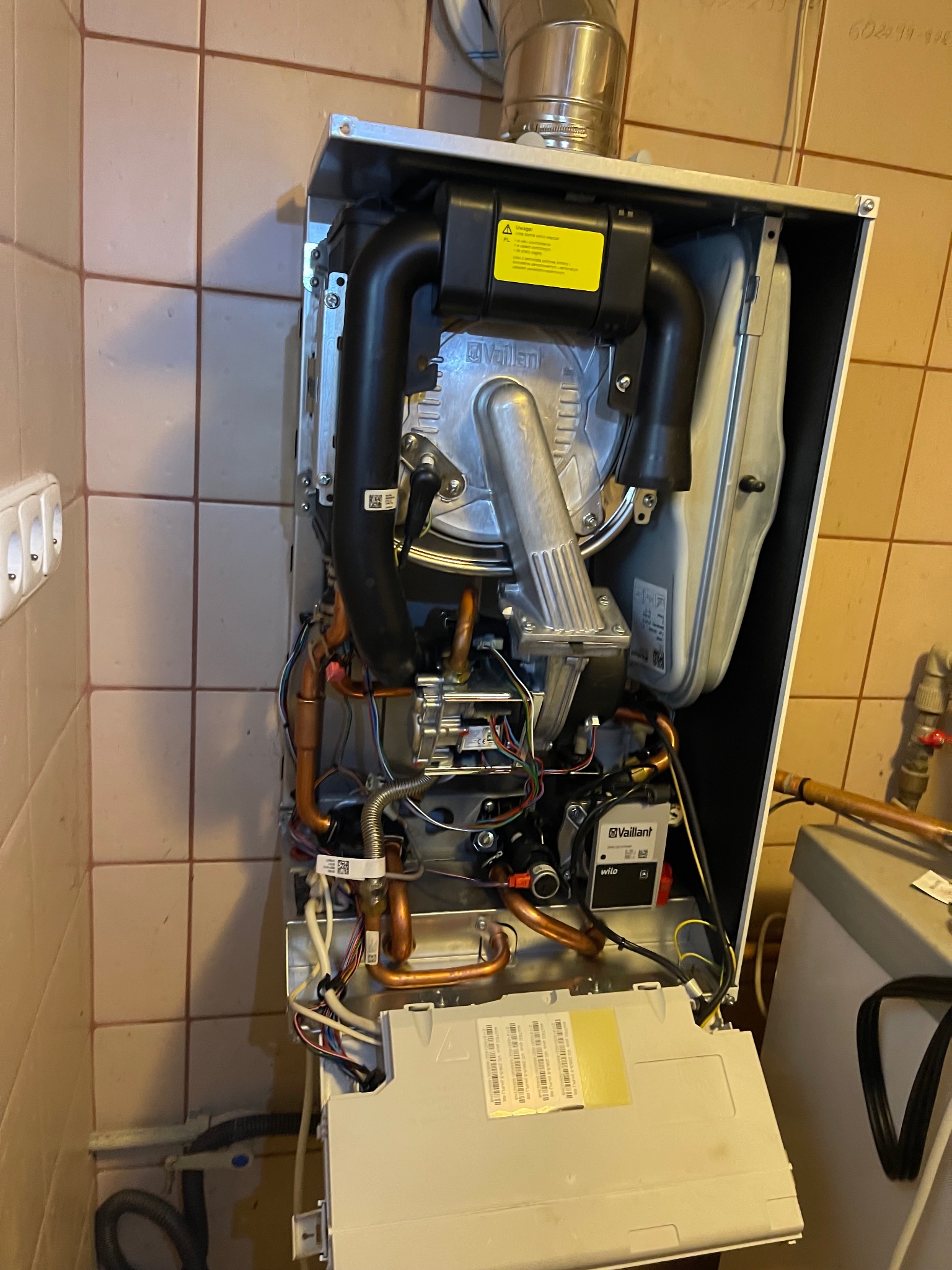 Otwarty piec gazowy Vaillant z widocznymi elementami instalacji, rurami miedzianymi i okablowaniem, na tle beżowych płytek. Widoczne przyłącza i osprzęt.