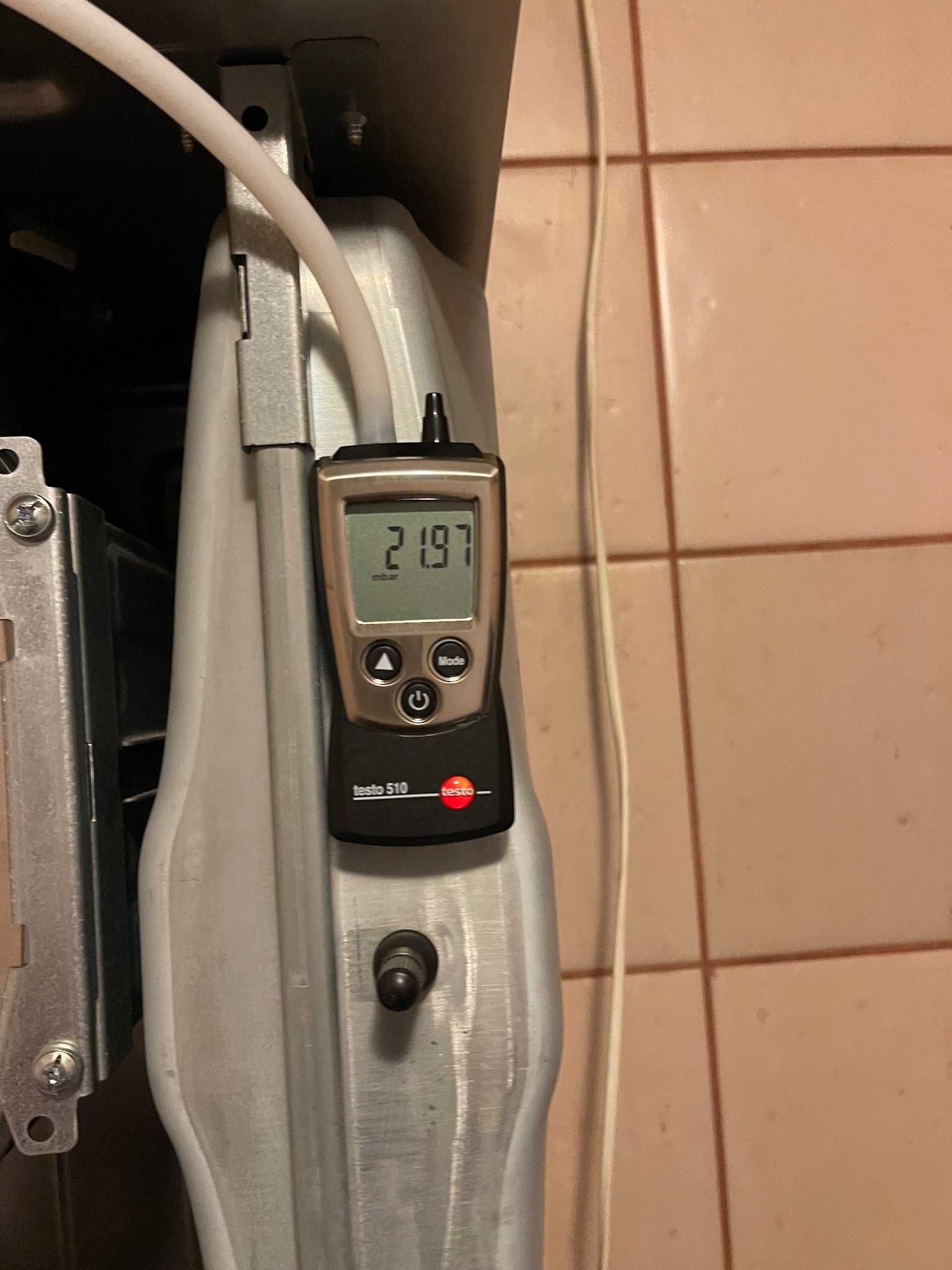 Miernik ciśnienia gazu Testo 510 przy piecu gazowym, wskazujący odczyt 21.97 mbar, podczas serwisu. Widoczne elementy instalacji i płytki ceramiczne w tle.