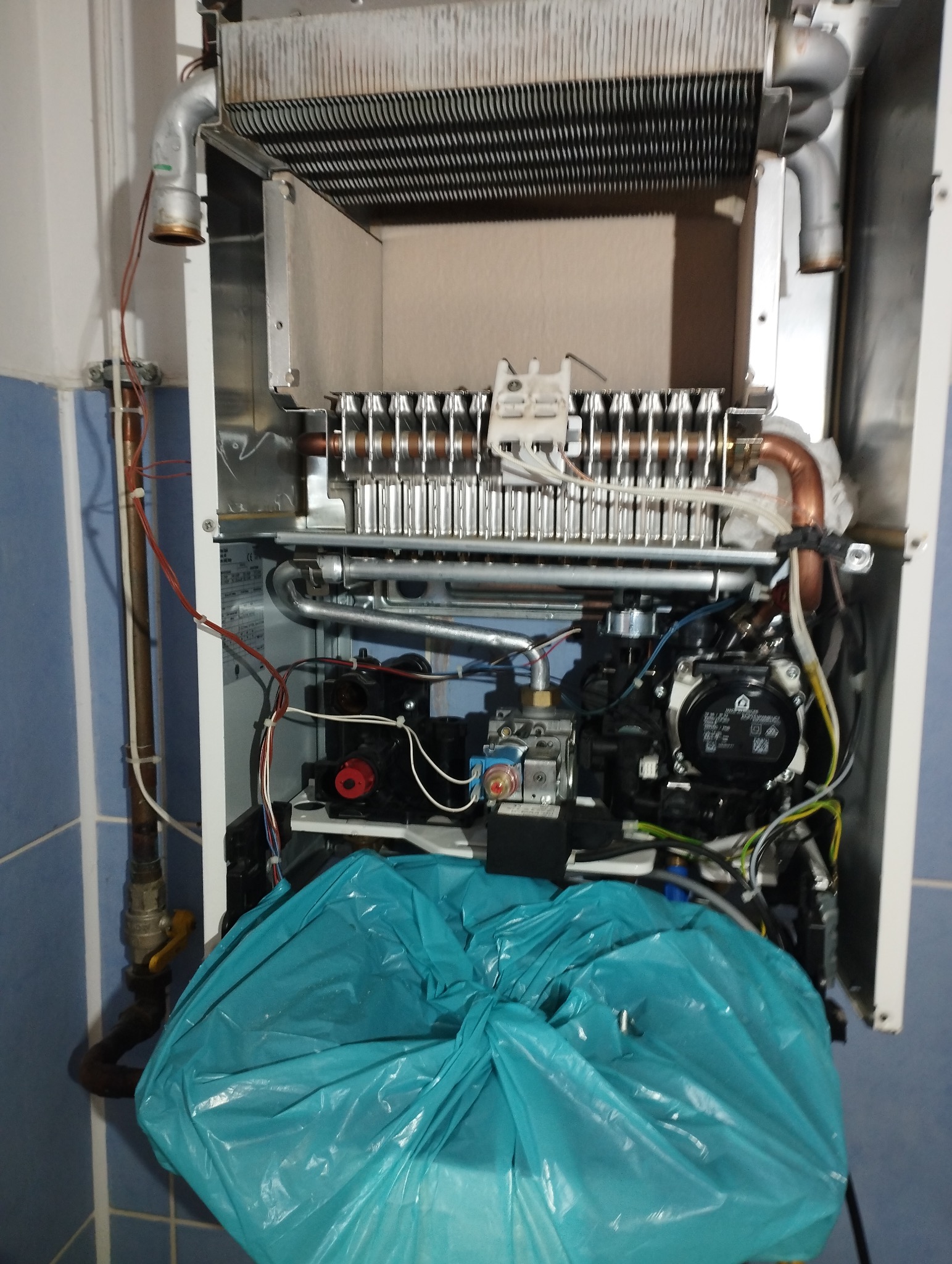 Otwarty piec gazowy z widocznymi elementami wewnętrznymi, zabezpieczony folią. Detal kotła c.o. w trakcie serwisu, z elementami instalacji i przewodami.