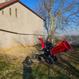 Czerwony rębak do gałęzi marki Cedrus stoi na trawniku obok budynku z jasną elewacją i stertą zrębków, w tle widoczne drzewo bez liści i ogrodzenie.