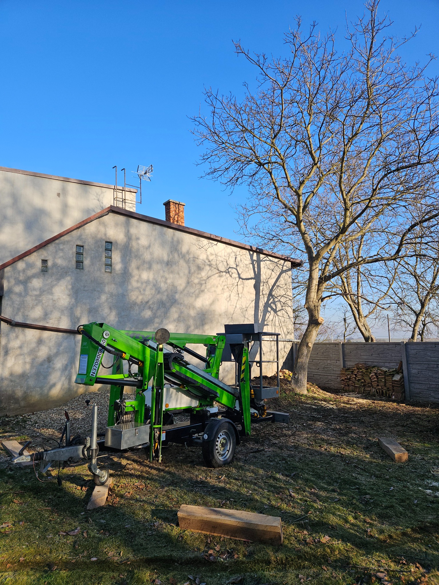 Zielony podnośnik koszowy na przyczepie, przygotowany do pracy przy drzewie z obciętymi gałęziami, na tle budynku i błękitnego nieba.