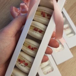 DOMOWA PRACOWNIA CUKIERNICZA WYPIECZONE Natalia Siadul - Ręka trzyma białe pudełko z makaronikami o smaku cytrynowym z dodatkiem malin, przewiązane różową wstążką. W tle widoczne kolejne pudełka z makaronikami.
