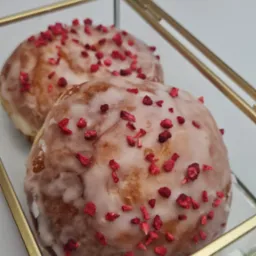 Dwa pączki z lukrem i posypką z liofilizowanych malin, ułożone w szklanym pudełku z pozłacanymi brzegami, na białym tle.