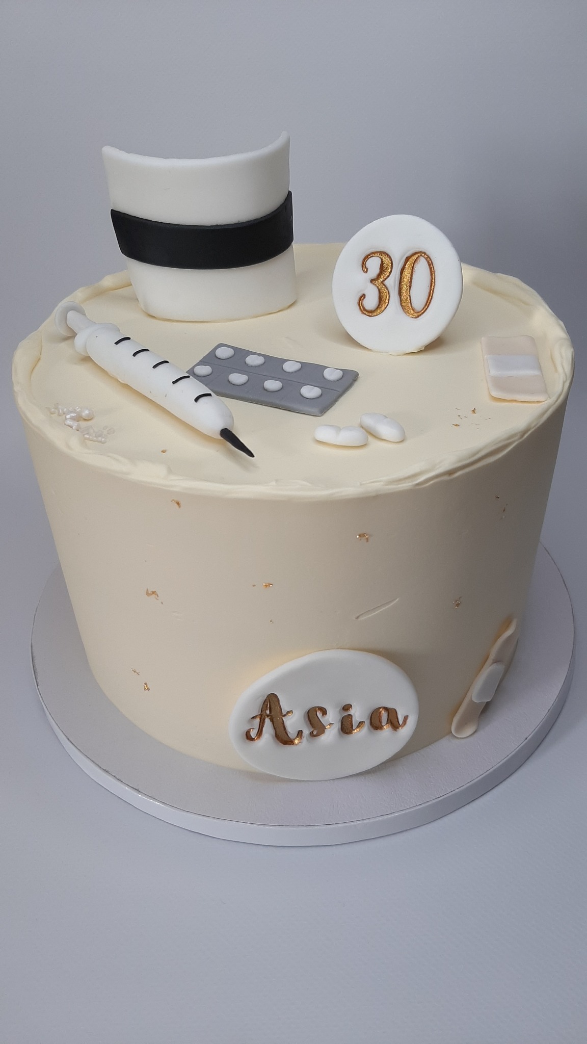 Tort urodzinowy z motywem medycznym: czapka pielęgniarki, strzykawka, tabletki i plasterki z napisem 'Asia' wykonanym złotą czcionką.