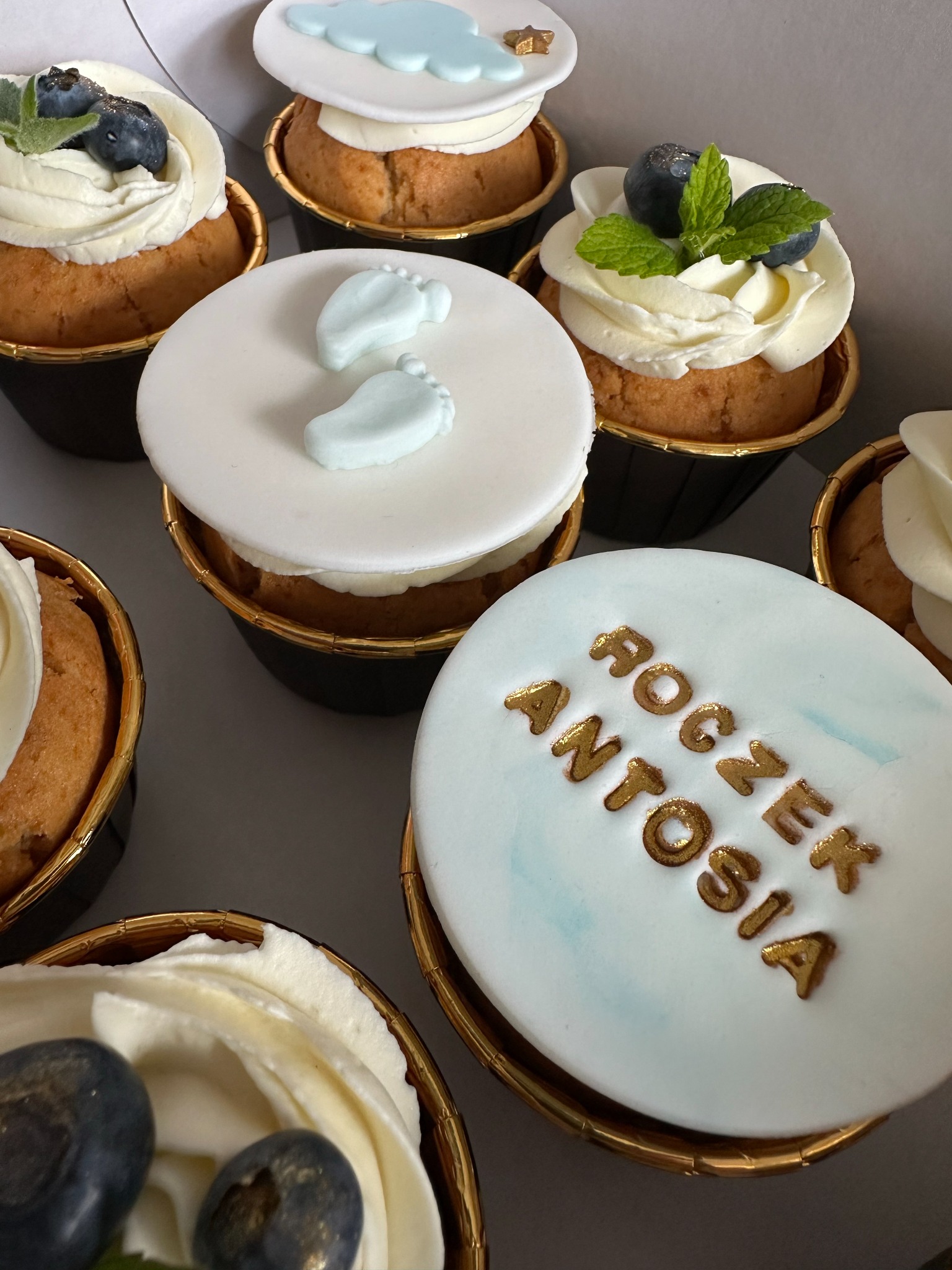 Dekorowane babeczki w papilotkach z motywami dziecięcymi: biały lukier z odciśniętymi złotymi literami 'Roczek Antosia', błękitne stópki i chmurki, krem z borówkami i miętą.