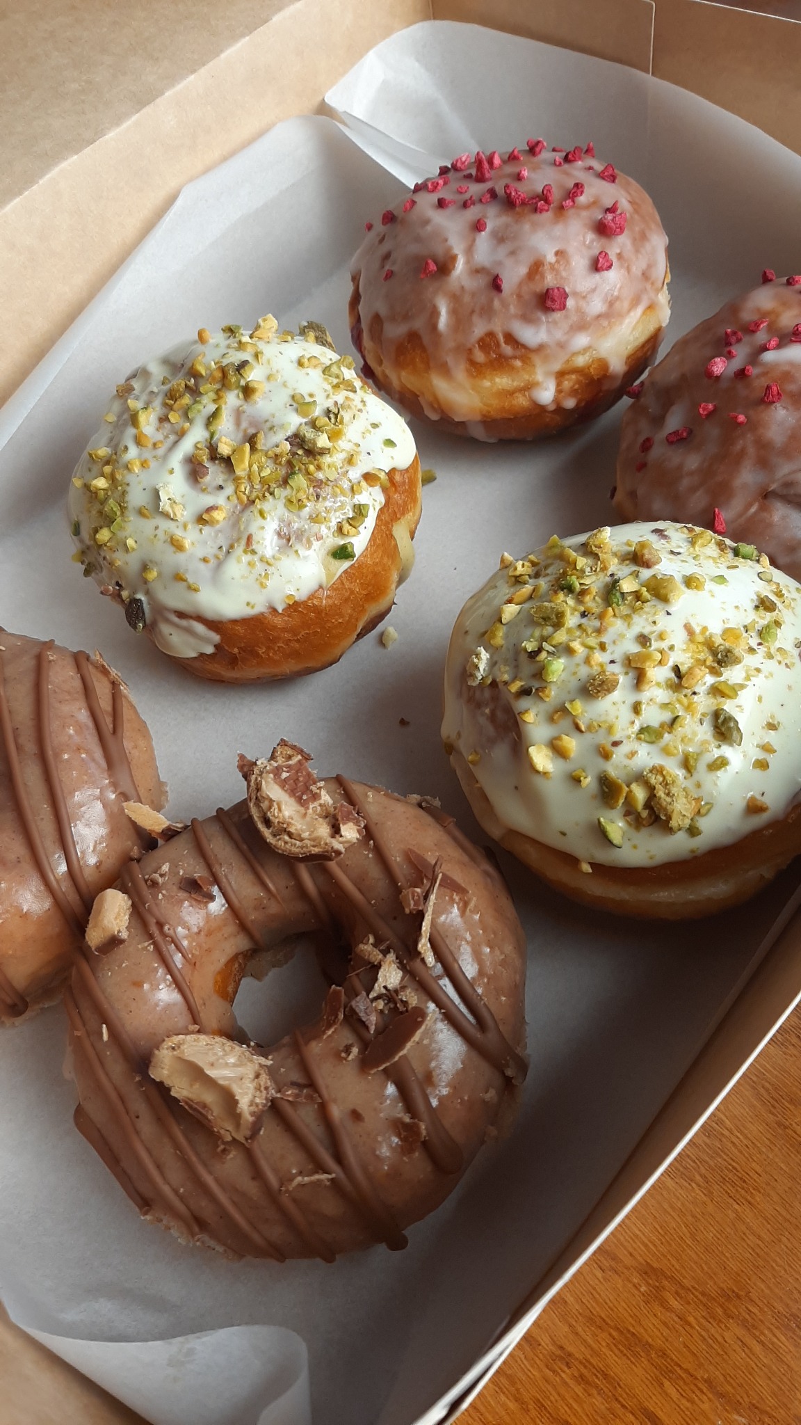 Różnorodne pączki w kartonowym pudełku: dwa z polewą pistacjową, dwa z różową posypką i dwa z polewą czekoladową, jeden z kawałkiem batonika na wierzchu.