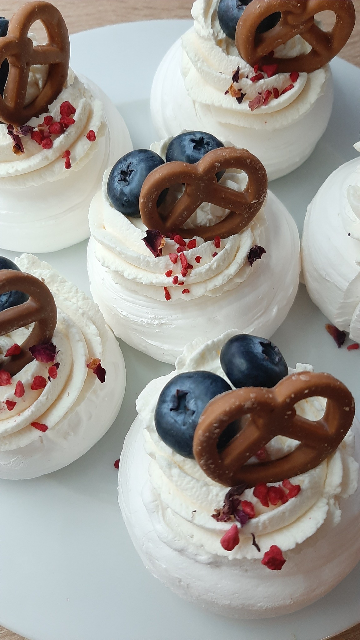 Mini bezy z bitą śmietaną, borówkami, precelkami i posypką z suszonych płatków róż i malin, ułożone na białym talerzu.