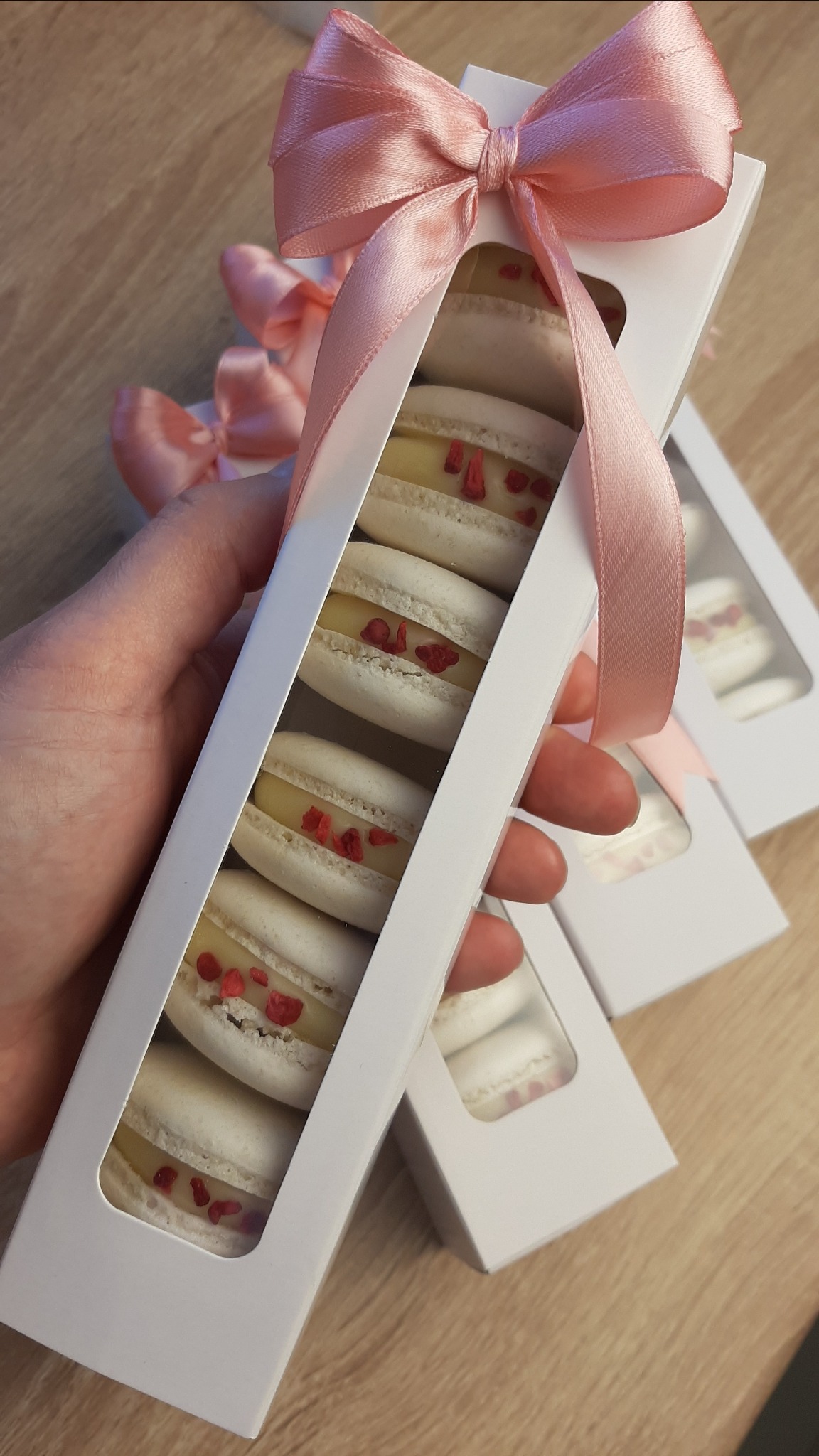 Ręka trzyma białe pudełko z makaronikami o smaku cytrynowym z dodatkiem malin, przewiązane różową wstążką. W tle widoczne kolejne pudełka z makaronikami.