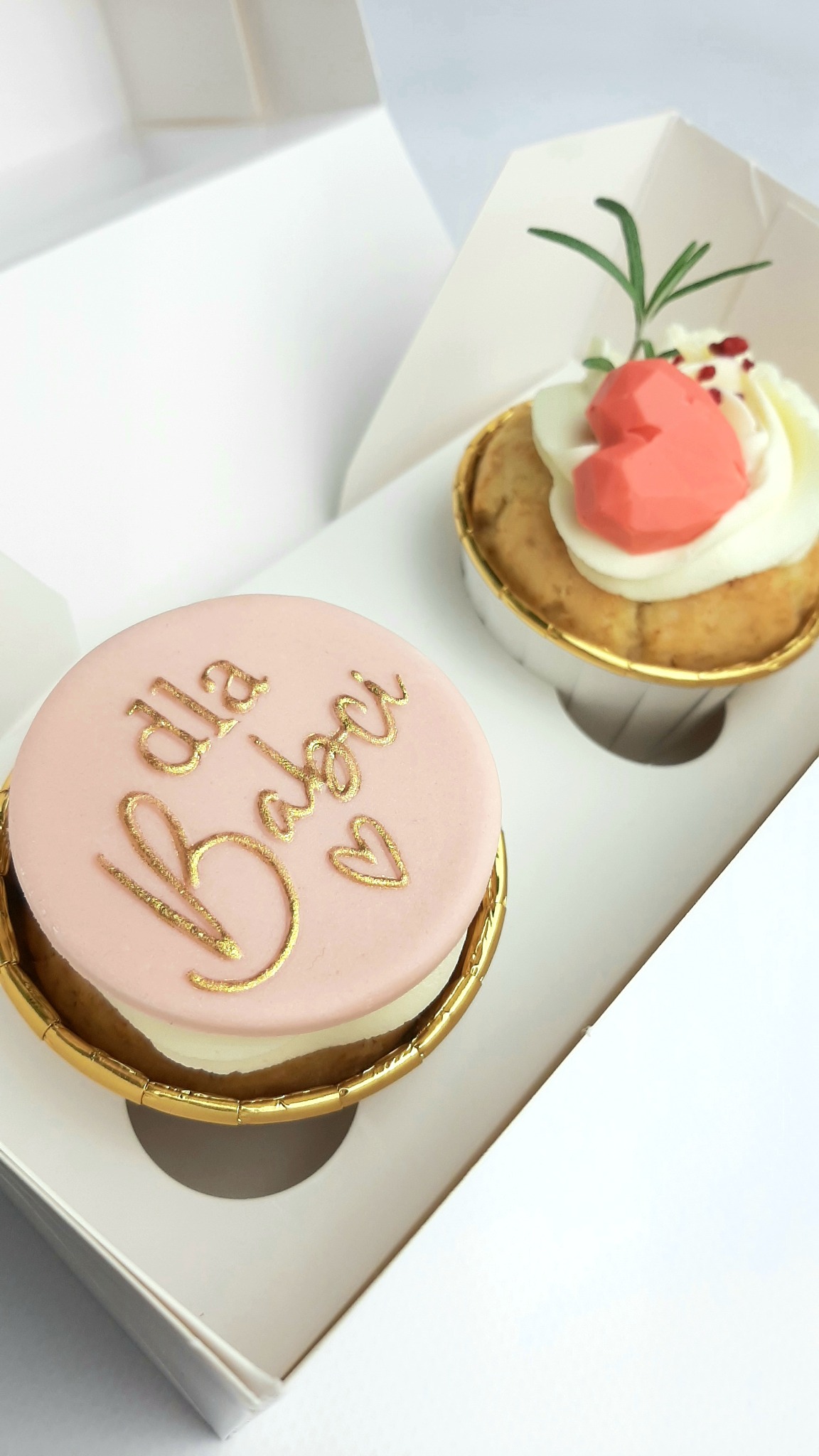 Dwa ozdobne babeczki w białym pudełku. Pierwsza z różowym lukrem i złotym napisem 'Dla Babci' oraz serduszkiem, druga z białym kremem, różowym sercem z czekolady i gałązką rozmarynu.