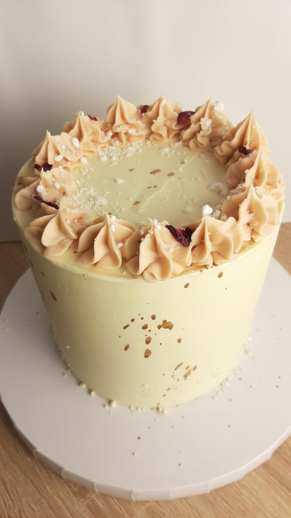 Tort z kremowym lukrem w odcieniach bieli i beżu, udekorowany rozetkami z kremu, posypany białymi okruchami i płatkami róż, na drewnianym blacie.