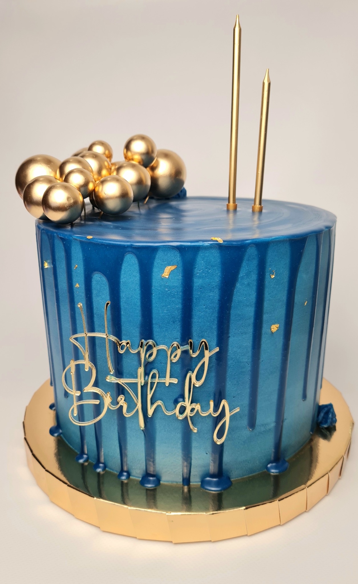 Niebieski tort urodzinowy z polewą, złotymi kulkami, napisem 'Happy Birthday' i dwiema świeczkami na złotym podkładzie.