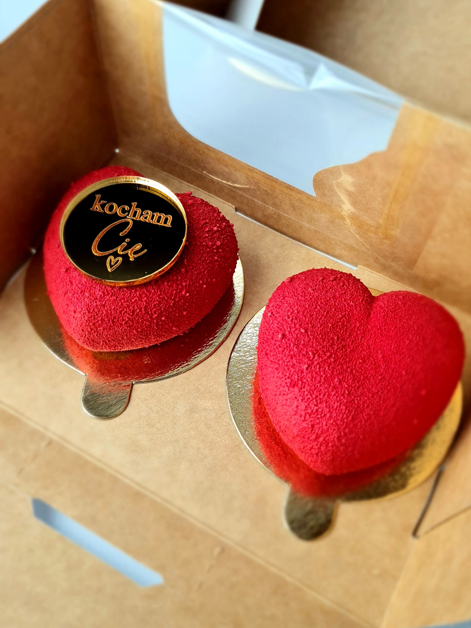 Dwa czerwone ciastka w kształcie serca na złotych podkładkach w kartonowym pudełku; jedno z czarnym, okrągłym topperem z napisem 'kocham Cię' w złotym kolorze.