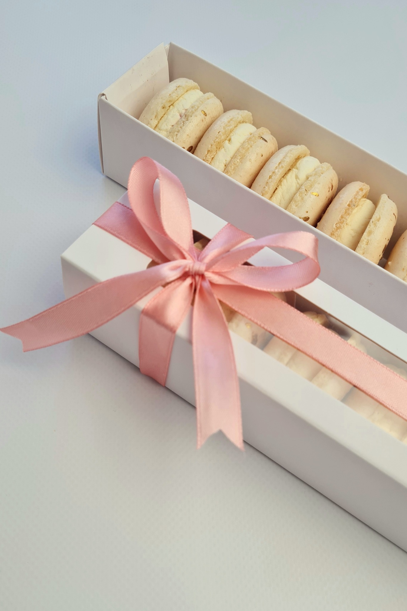 Delikatne, jasne makaroniki z kremowym nadzieniem, z widocznymi drobinkami złota, ułożone w białym pudełku z okienkiem, przewiązanym różową satynową wstążką.