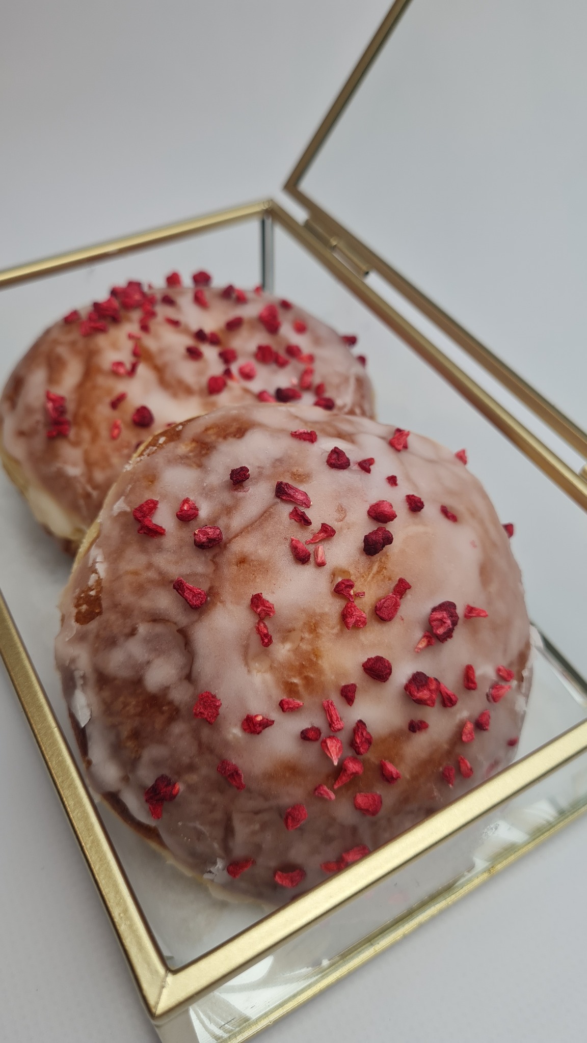 Dwa pączki z lukrem i posypką z liofilizowanych malin, ułożone w szklanym pudełku z pozłacanymi brzegami, na białym tle.