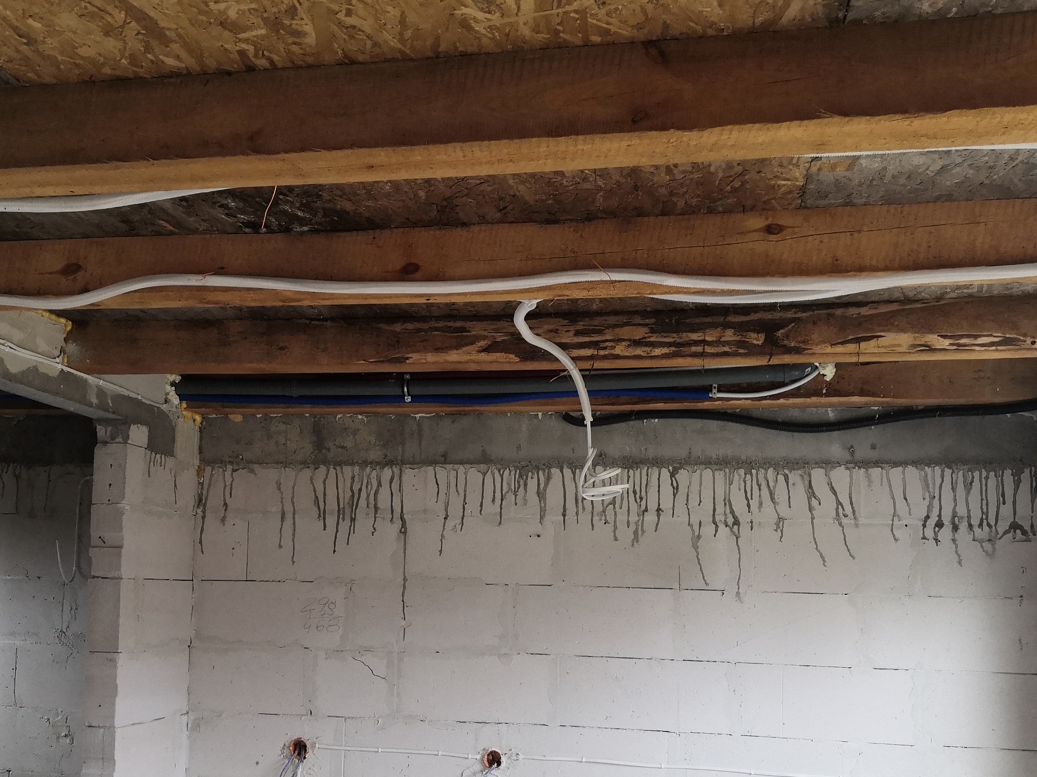 Wewnętrzna instalacja przewodów elektrycznych i rur pod stropem drewnianym w surowym stanie, widoczne białe kable, niebieskie rury oraz szare ściany z betonu komórkowego z zaciekami.