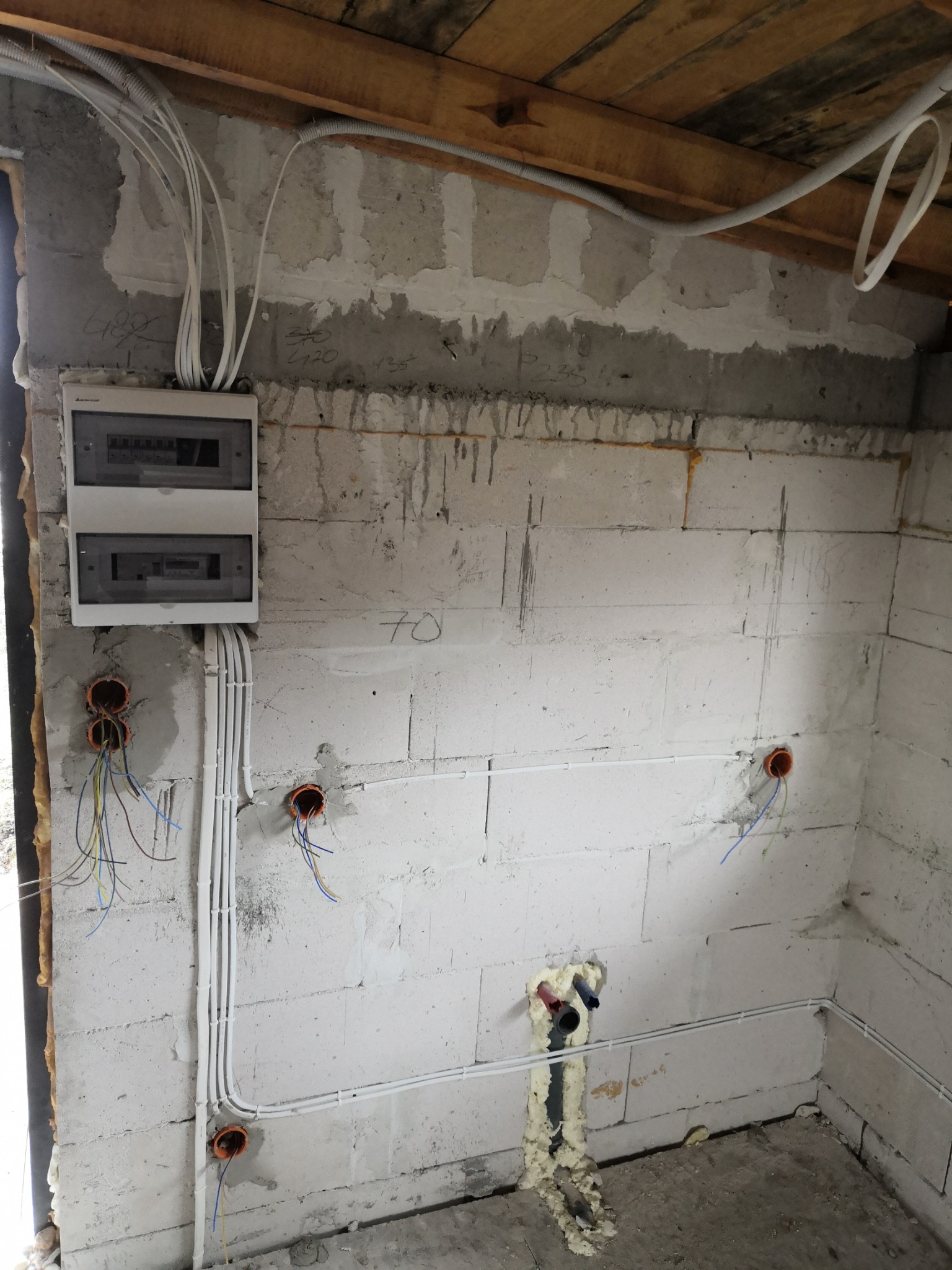 Rozdzielnia elektryczna z poprowadzonymi przewodami w białych peszlach, widoczne otwory w ścianie z wystającymi kablami, ściana z betonu komórkowego w stanie surowym.