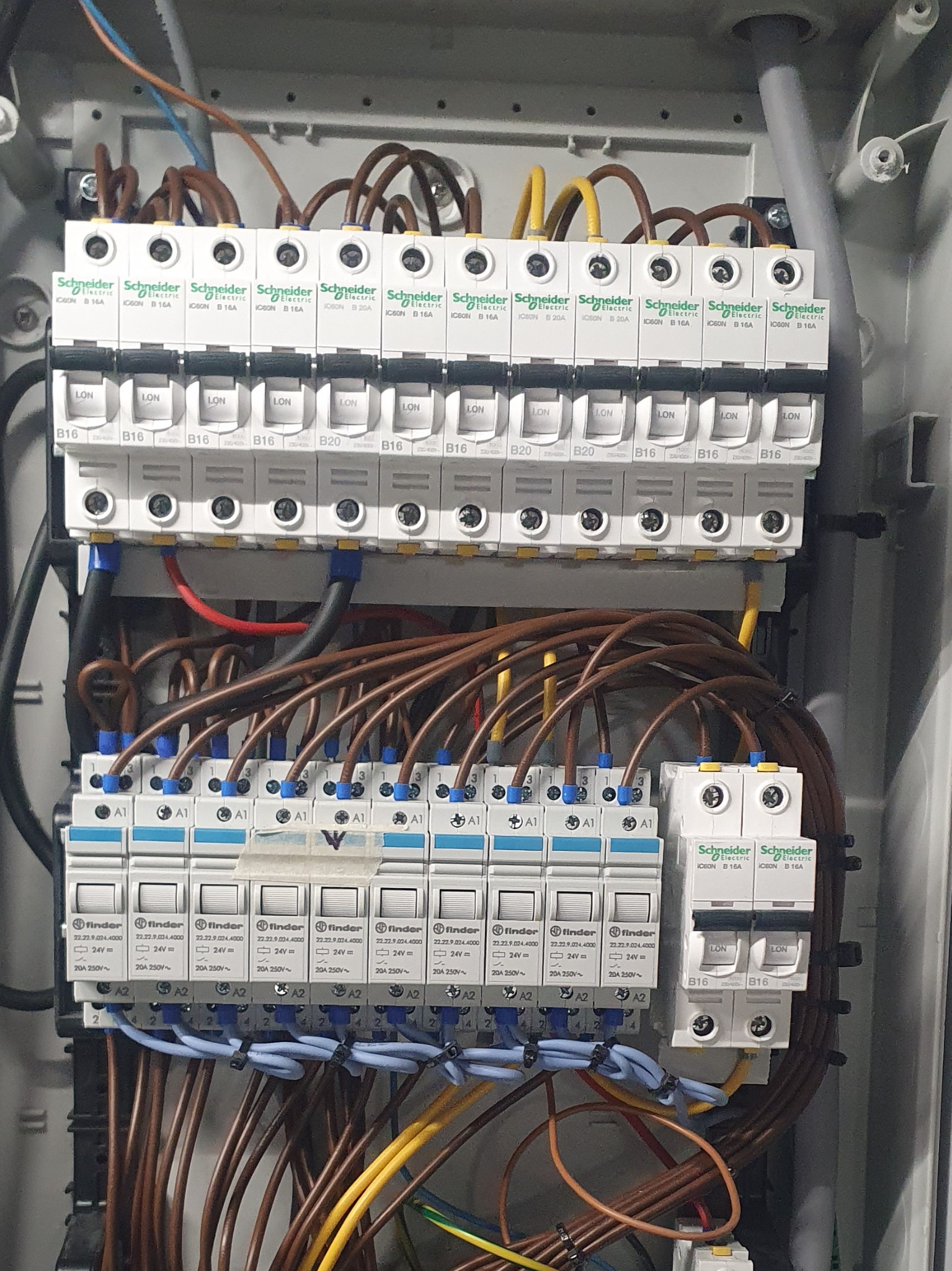 Rozdzielnica elektryczna z zainstalowanymi bezpiecznikami i przekaźnikami różnych producentów (Schneider Electric, Finder), widoczne brązowe, żółte i niebieskie przewody, zasilanie 24V...