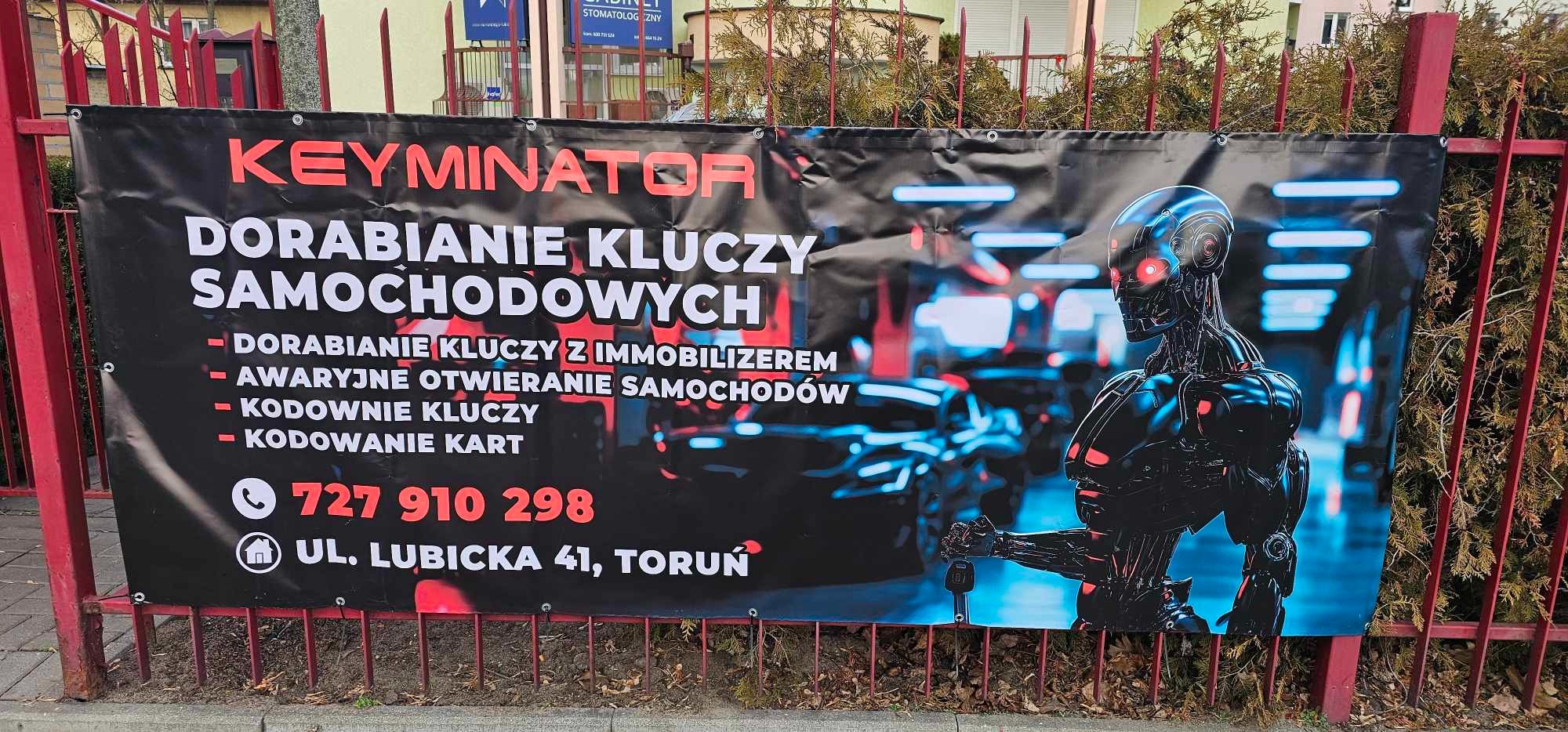 Baner reklamowy firmy Keyminator oferującej dorabianie kluczy samochodowych, awaryjne otwieranie samochodów i kodowanie kart, z futurystyczną grafiką robota trzymającego kluczyk w dłoni, zawieszony...