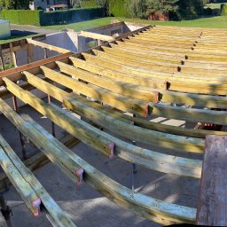 LUXHOUSE - Usługi budowlane Mariusz Kryger - Wysoki kąt: drewniane belki stropowe z czerwonymi łącznikami, wsparte na metalowych podporach, w surowym stanie, na tle zieleni i częściowo wymurowanych ścian.