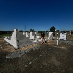 LUXHOUSE - Usługi budowlane Mariusz Kryger - Budowa domu z bloczków Solbet w słoneczny dzień. Widoczni pracownicy i częściowo wzniesione ściany fundamentowe. Na drugim planie zapakowane bloczki i tyczka geodezyjna.