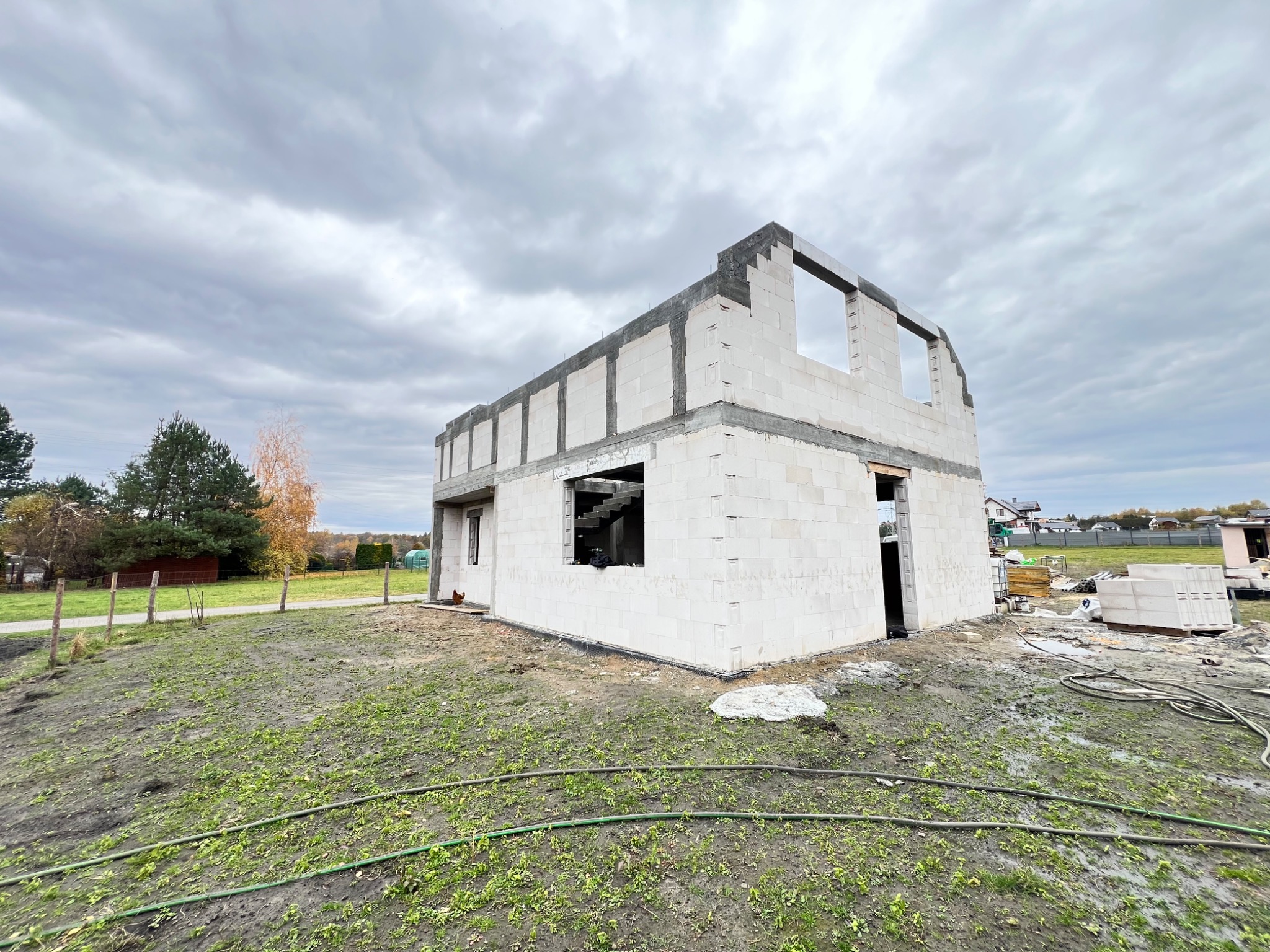 Surowy stan dwukondygnacyjnego domu z białego betonu komórkowego, z widocznymi otworami okiennymi i drzwiami, na tle pochmurnego nieba i zielonego terenu.