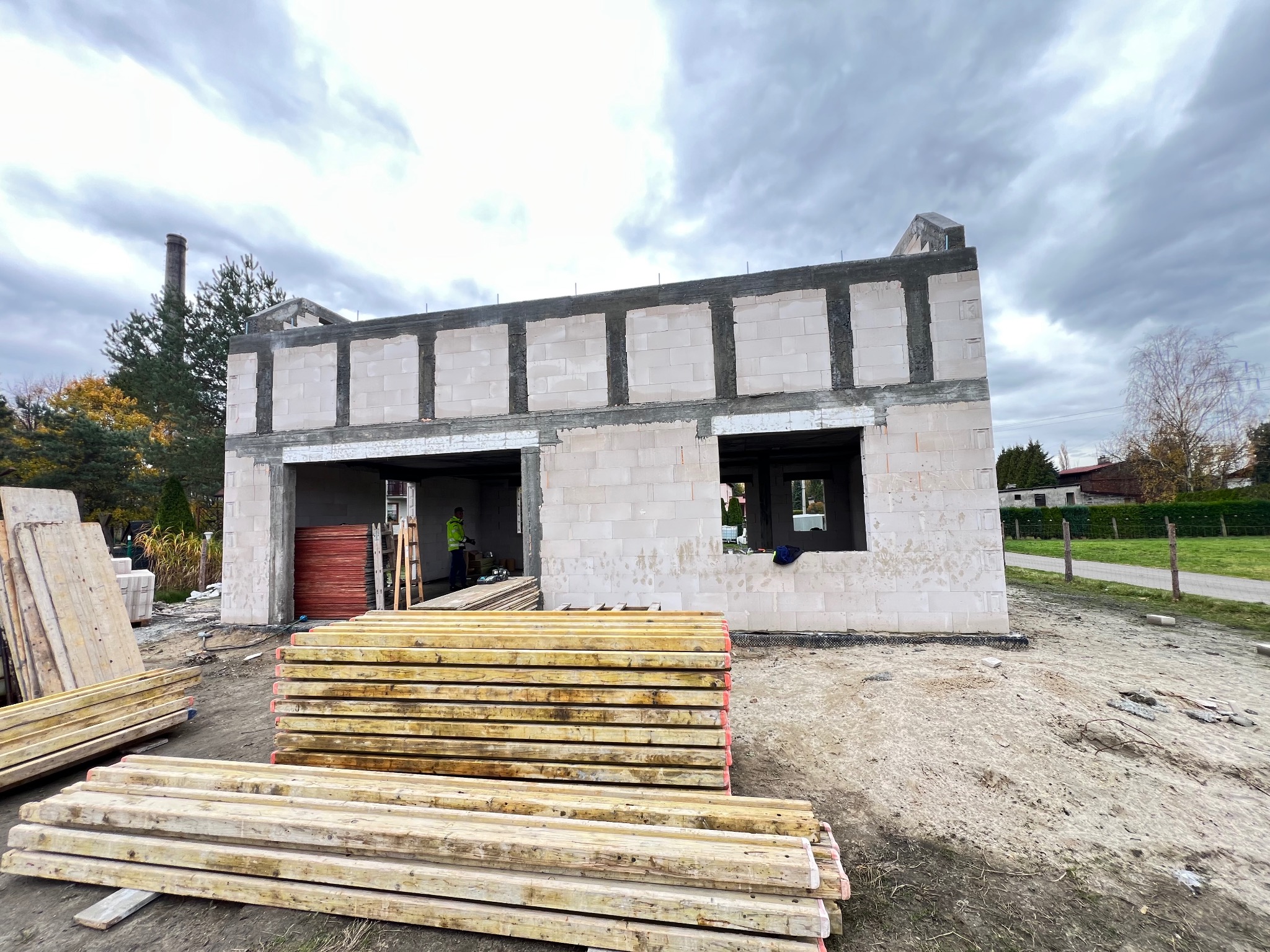 Budowa domu w trakcie: ściany z betonu komórkowego, widoczne szalunki i deski. W tle drzewa i pochmurne niebo. Etap murowania ścian.