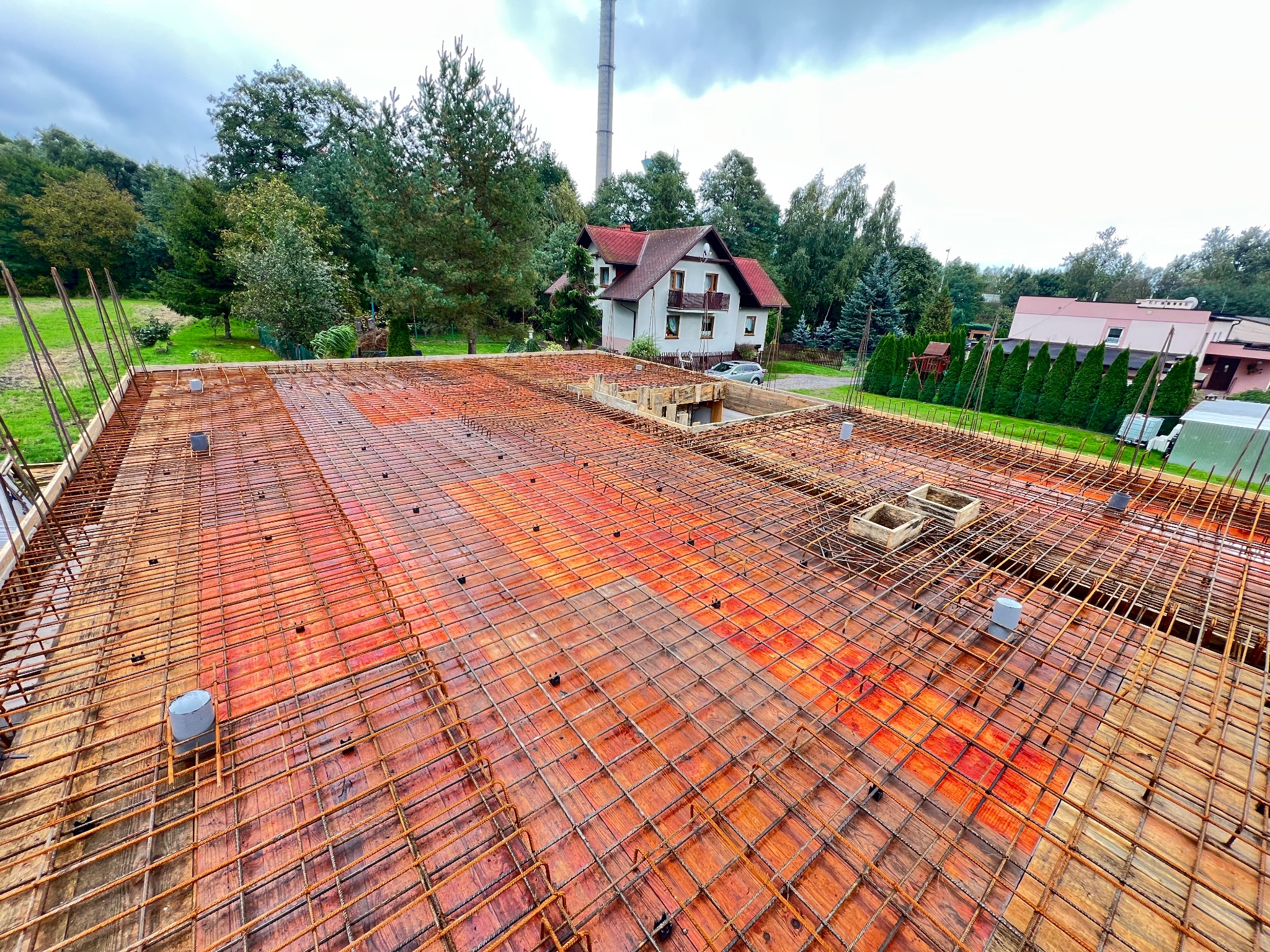 Zbrojenie stropu domu jednorodzinnego z widoczną siatką z prętów stalowych, szalunkiem i elementami instalacji. W tle dom i komin.