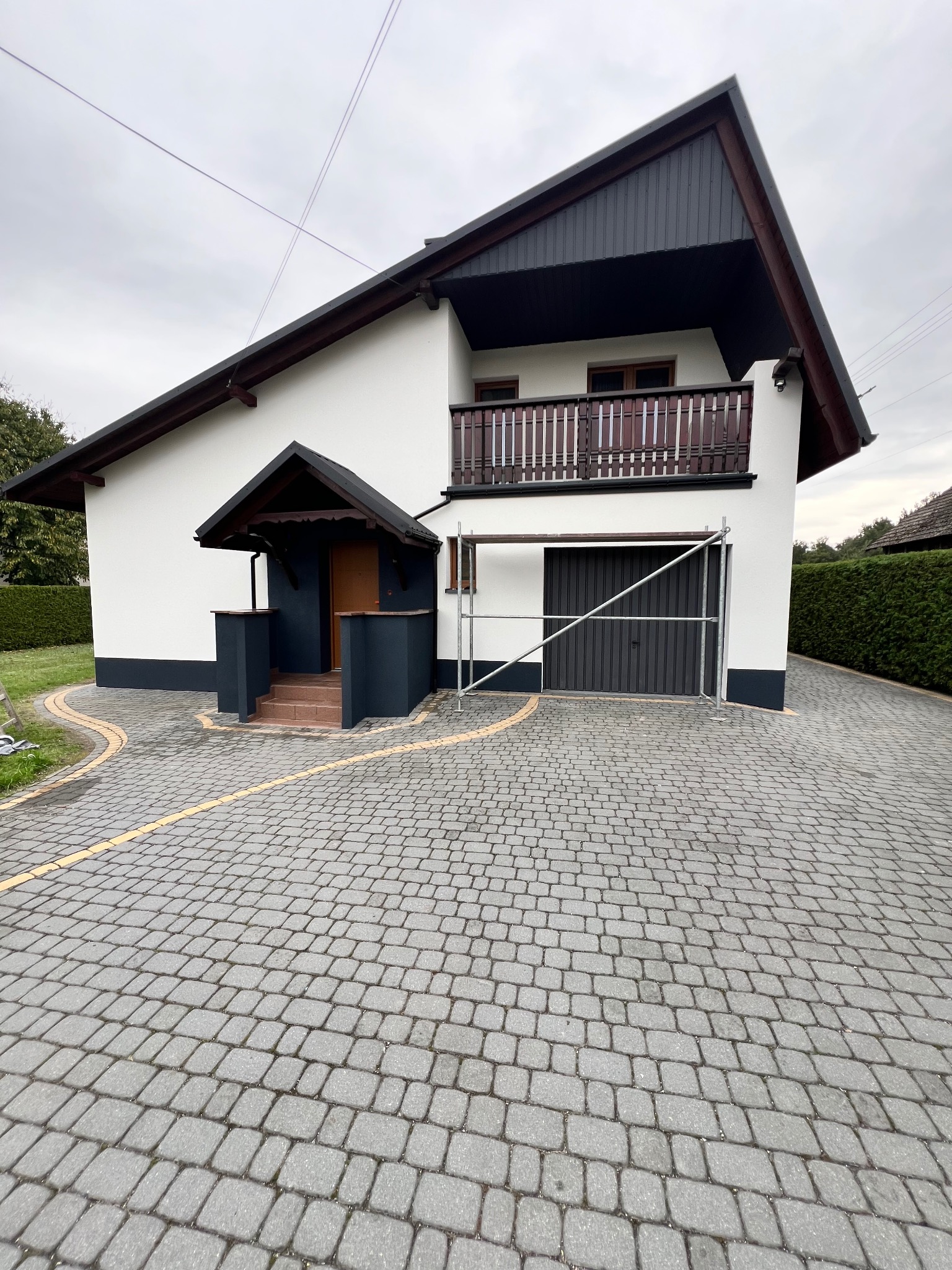 Dwukondygnacyjny dom z balkonem i garażem, po malowaniu elewacji na biało i grafitowo. Widoczne rusztowanie przy garażu. Podjazd wyłożony kostką brukową.