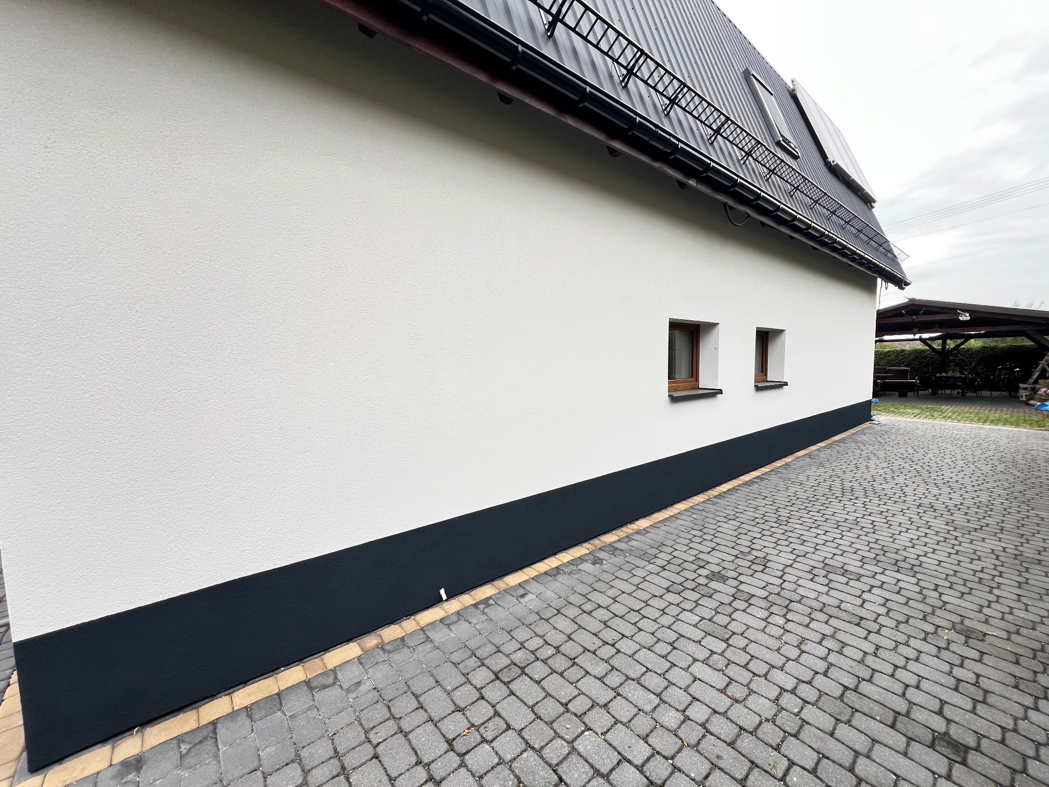 Boczny widok domu z białą elewacją i ciemnym cokołem, małe okna, szara kostka brukowa na podjeździe, metalowe pokrycie dachu, pochmurne niebo.