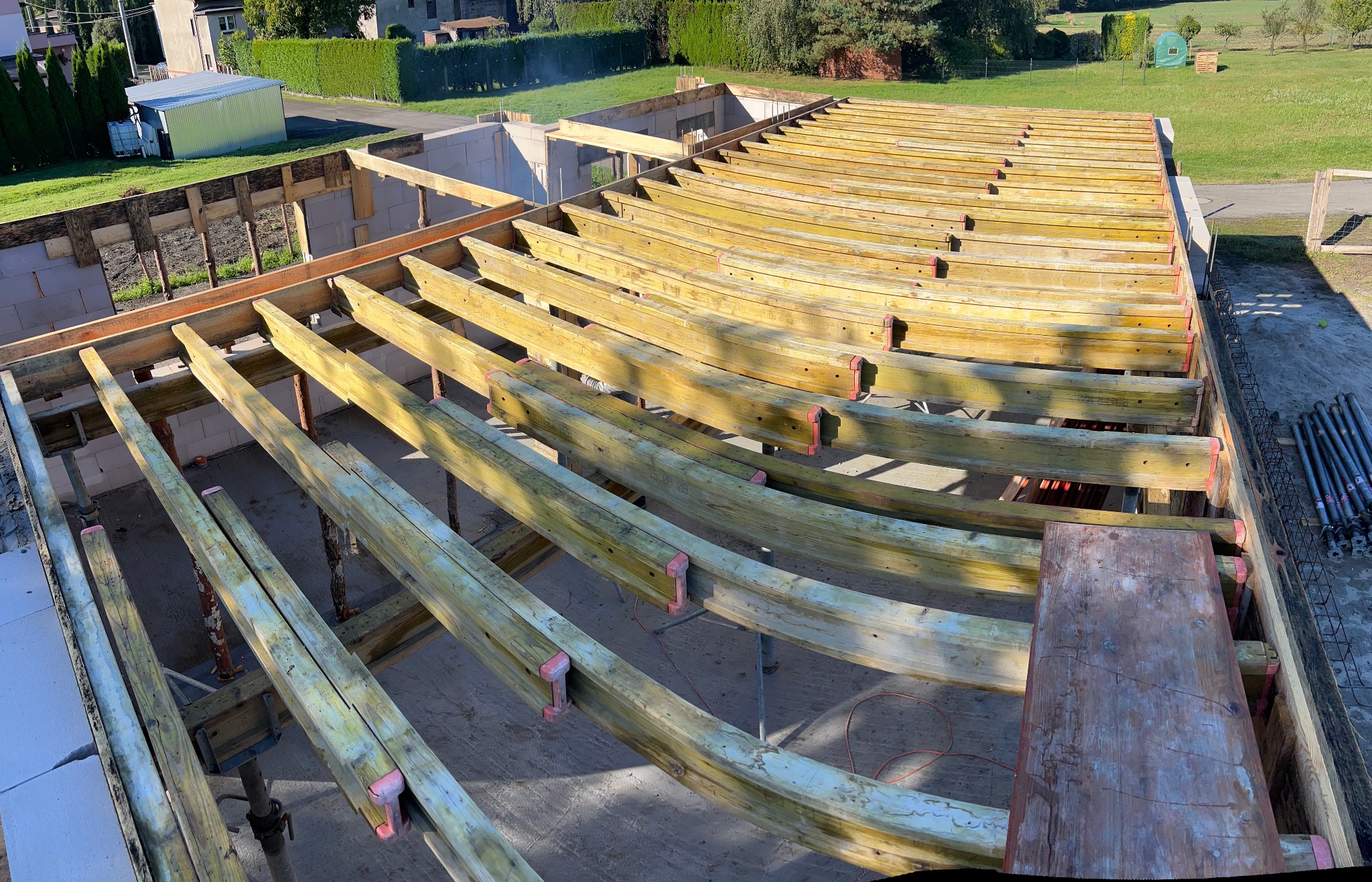 Wysoki kąt: drewniane belki stropowe z czerwonymi łącznikami, wsparte na metalowych podporach, w surowym stanie, na tle zieleni i częściowo wymurowanych ścian.