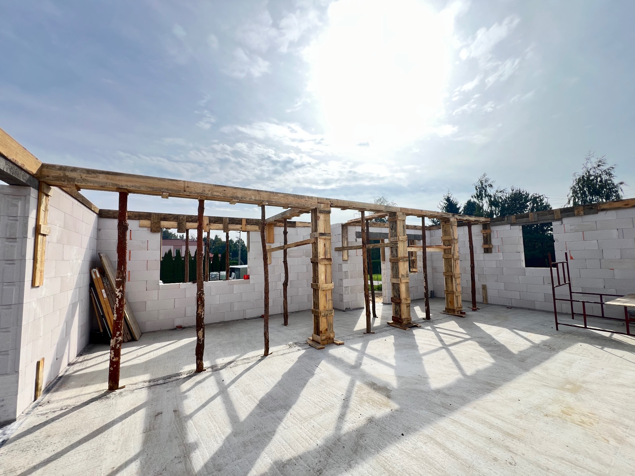 Wnętrze surowego domu z betonu komórkowego w trakcie budowy. Widoczne drewniane stemple podtrzymujące strop i ściany z pustaków. Słoneczny dzień.