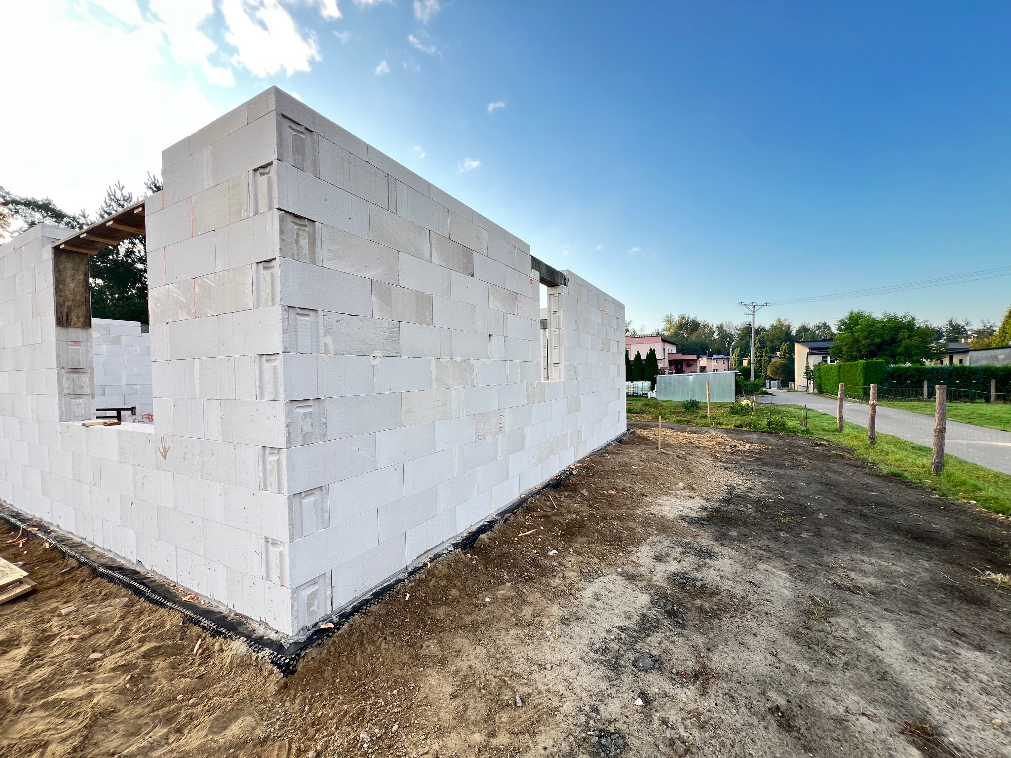 Wznoszenie ścian domu z białych bloczków, widoczne otwory okienne i drzwiowe. Na pierwszym planie grunt, w tle zabudowa jednorodzinna i błękitne niebo.