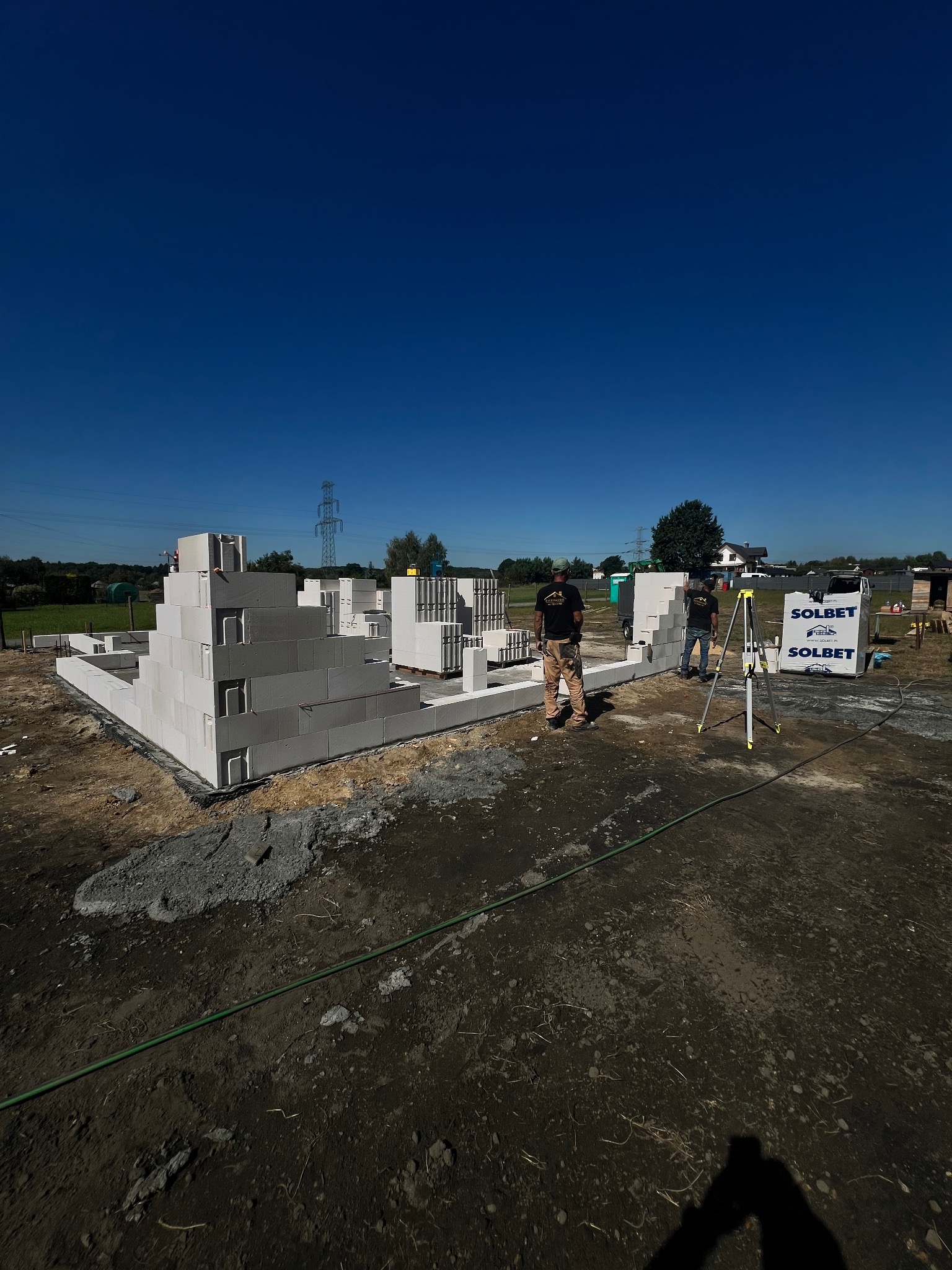 Budowa domu z bloczków Solbet w słoneczny dzień. Widoczni pracownicy i częściowo wzniesione ściany fundamentowe. Na drugim planie zapakowane bloczki i tyczka geodezyjna.