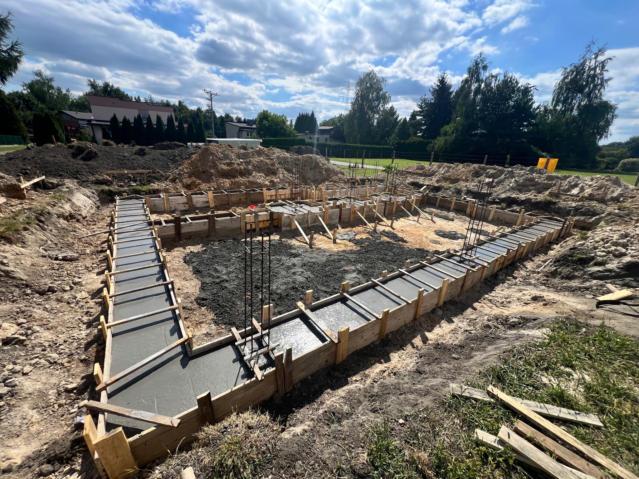 Zalane betonem fundamenty domu w drewnianym szalunku, widoczne zbrojenia. W tle ziemia, drzewa i błękitne niebo z chmurami. Ujęcie z góry, szeroki kadr.